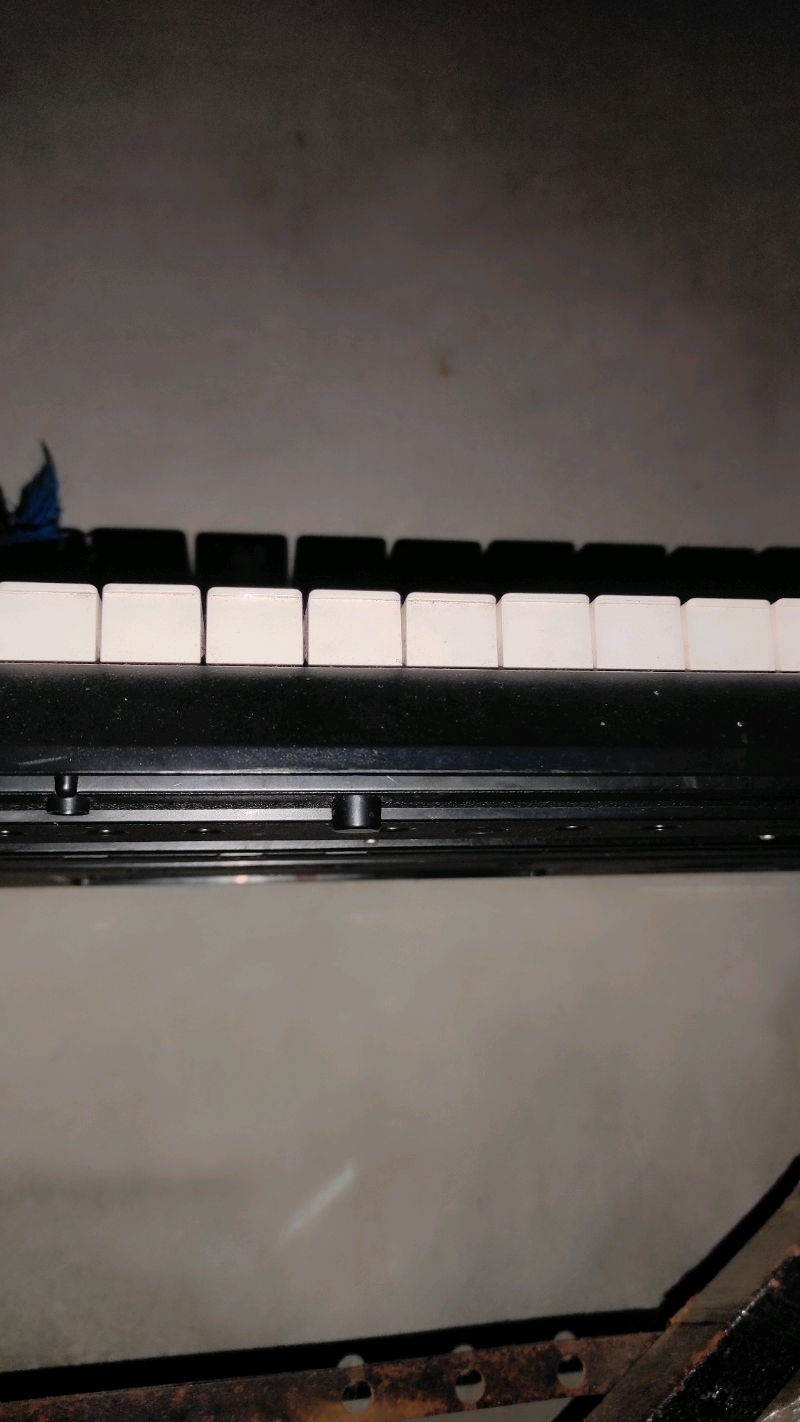 The image shows a close-up, horizontal, eye-level shot of a black keyboard with white keys. The keyboard is positioned in the foreground, with a dark, textured background that appears to be a wall or floor. The lighting is dim and appears to be artificial, casting shadows on the keys. There is a blue object partially visible on the left side of the keyboard, and some rust or dirt on the surface below the keyboard. The image does not contain any visible people or actions. The location in Guwosari, Indonesia is not discernible from the image itself.