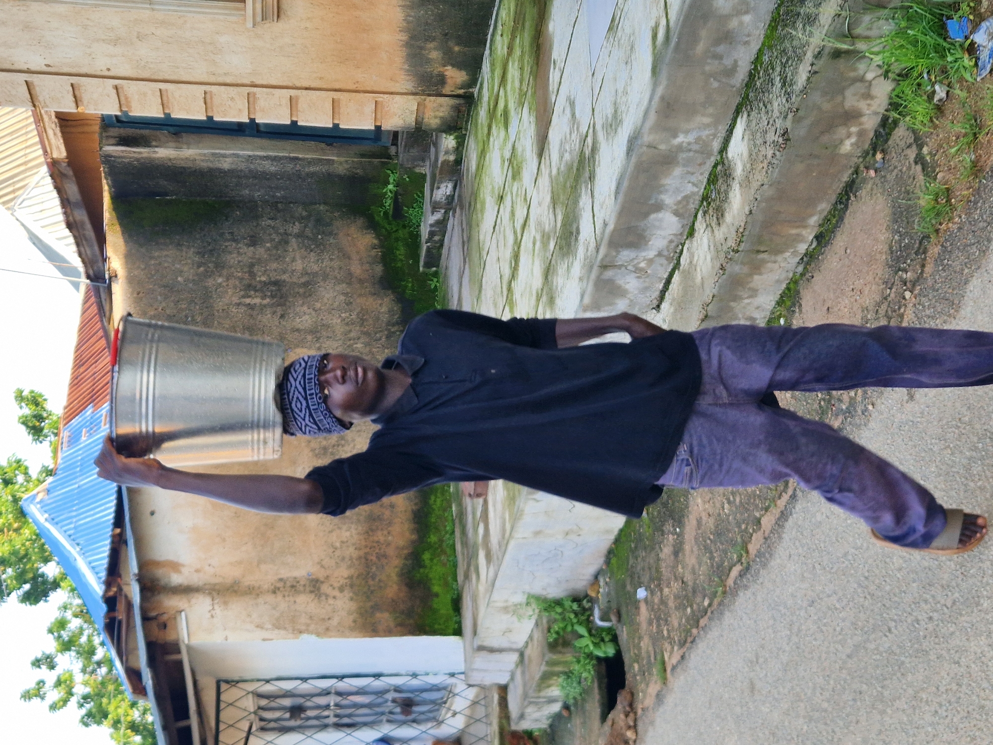 A young male is captured in a medium shot from the waist up, walking on a paved path outdoors. He is wearing a black collared shirt and purple pants. A metal bucket is balanced on his head, secured by a patterned fabric headband. His right hand is extended upwards, holding the bucket steady, while his left hand rests in his pocket. The background shows a building with a corrugated metal roof and a rough-textured wall. Green vegetation is visible along the path and on the building's structure. The ground is paved with concrete slabs, some of which are covered in moss. The scene suggests a typical day in Kabong, Nigeria, where such everyday tasks are common.