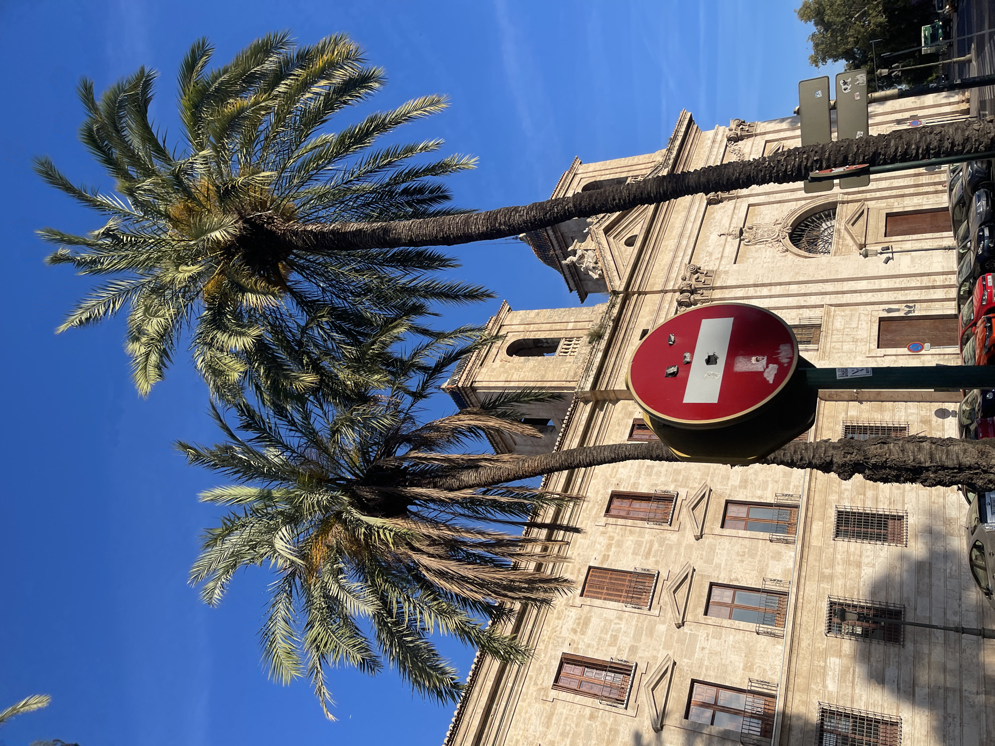 The image depicts a street scene in València, Spain, featuring two tall palm trees silhouetted against a clear blue sky and a light-colored, ornate building, possibly a church or government building. A red and white "no entry" traffic sign is prominently displayed, and the building's facade includes multiple windows with varying types of coverings and architectural details. Parked cars are visible on the right side of the frame, suggesting a street or avenue environment.