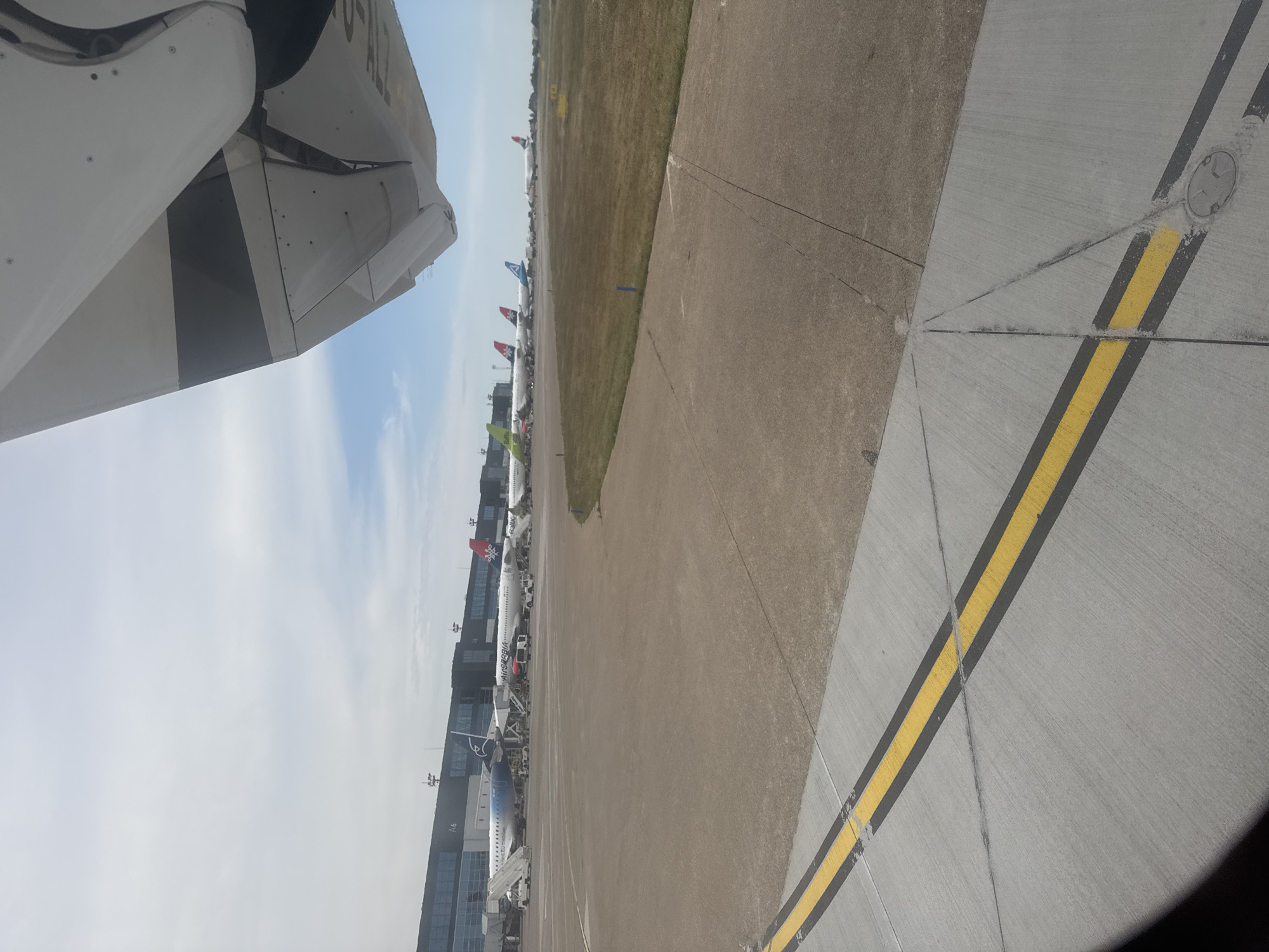 The image depicts an airport tarmac with several commercial airplanes lined up, likely at an air show or during a busy period. The scene is viewed from the perspective of someone on or near an aircraft, with a portion of its wing and fuselage visible in the foreground. The planes are of various airlines, indicated by their liveries and tail fins, some featuring prominent logos and colors. Flags are also visible atop some of the aircraft. In the background, an airport terminal building with multiple levels and windows stretches along the tarmac, suggesting an active airport environment. Adjacent to the tarmac is a grassy field, and beyond that, a hazy horizon. The sky is bright and appears to be partly cloudy, suggesting it is daytime, likely mid-morning or afternoon, with fair weather. The overall impression is one of an organized, bustling aviation setting. Some visible text on the planes includes what appears to be "AIR SERBIA" on one aircraft's fuselage and "A6" on the terminal building. The image is oriented vertically, requiring rotation for a standard landscape view.