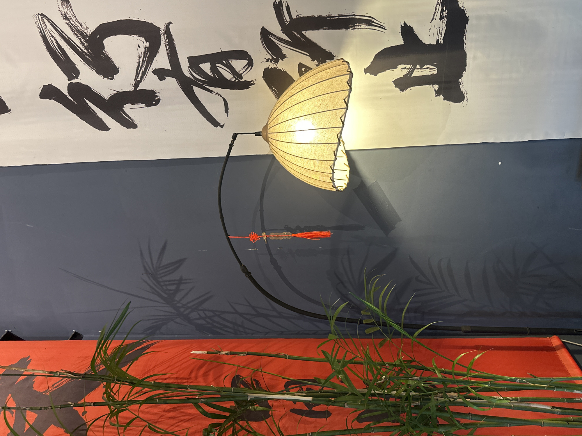 The image captures a scene inside a room in North Kuta, Indonesia. The background consists of a white wall with large black calligraphy characters on the upper half, and a gray wall below. A floor lamp with a lampshade that resembles a pleated fan is positioned in front of the wall. Attached to the lamp's arm is a red and gold ornamental tassel. The lower foreground is dominated by an orange surface with dark calligraphy characters, strewn with green bamboo stalks and leaves. The shadows of the bamboo leaves are cast on the gray wall.