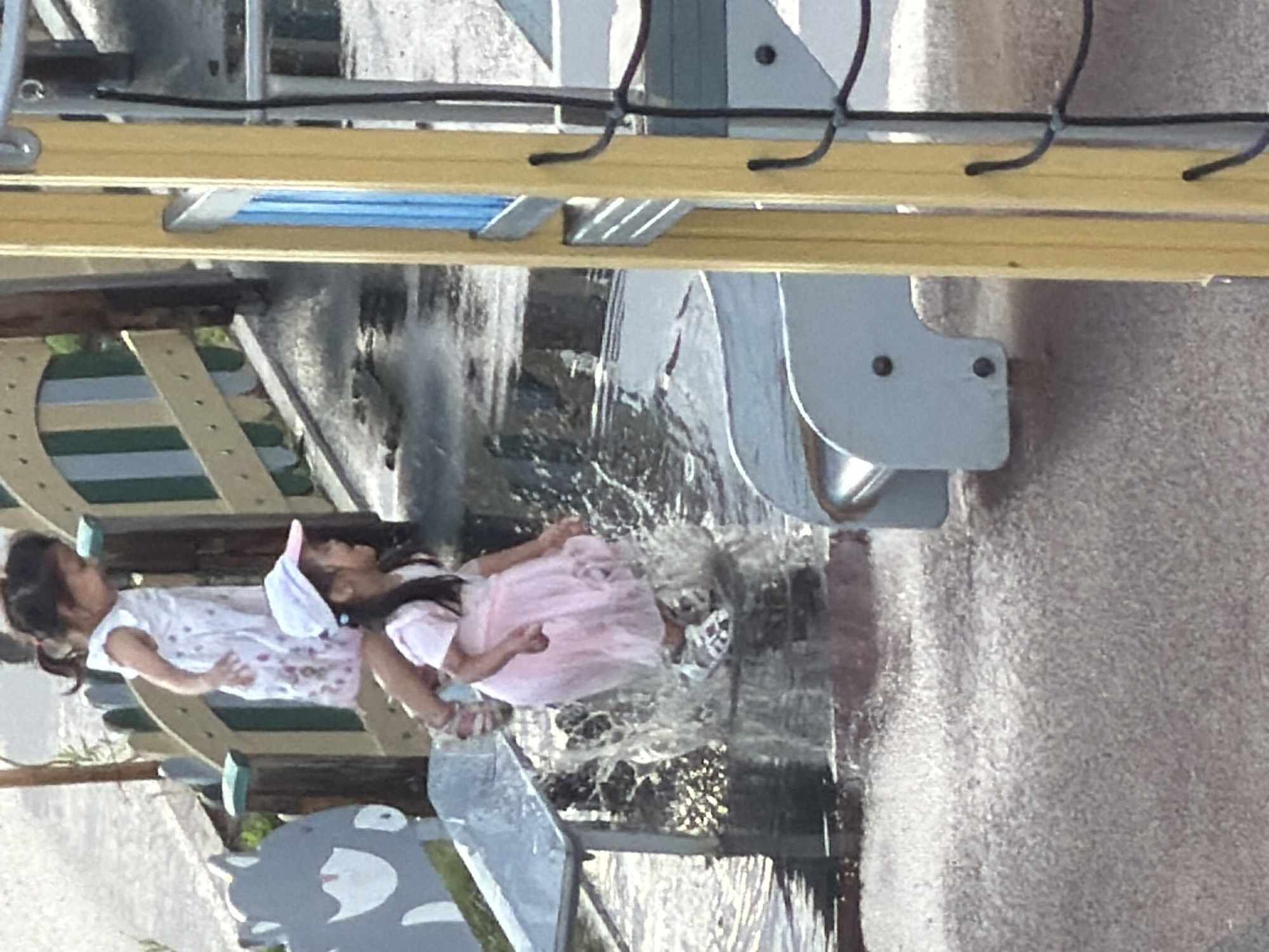 Two young girls are playing in a water feature, likely a splash pad, at a playground. One girl, wearing a pink dress and white sandals, is captured mid-jump, with water splashing around her. The other girl, wearing a pink top and a white cap, is standing to her left. The playground equipment includes a wooden structure with green and white striped panels and a gray, stylized animal-shaped decoration. The ground is wet and textured, appearing to be a type of rubberized surfacing. The scene is outdoors, with natural light illuminating the area. The image's composition suggests it was taken from a slightly elevated perspective, possibly by someone standing on a higher part of the playground or adjacent structure.