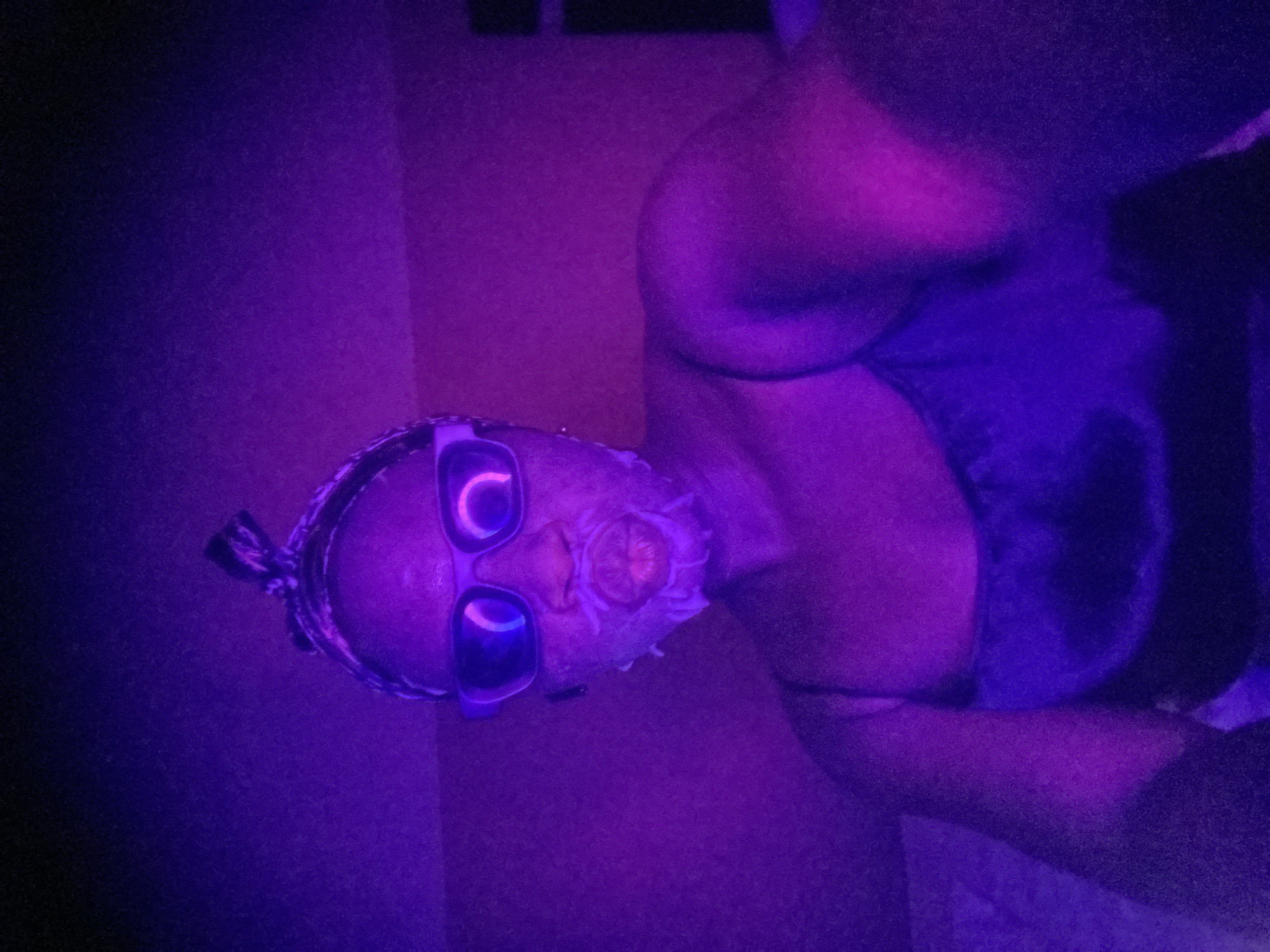 This sideways-oriented image features a person's upper body bathed in intense purple and blue artificial light, suggesting a black light or club environment. The subject has dark skin and is wearing glasses with light-colored frames that glow brightly under the purple illumination. Braided hair is visible towards the top of the head.
The person's face is adorned with distinctive white or light-colored makeup, possibly glitter or face paint, concentrated around the nose and mouth, which stands out prominently in the UV light. The lips are also highlighted in a similar light shade and are slightly parted. The visible clothing includes a dark top with broad straps, appearing on the torso.
The background is dark and indistinct, primarily consisting of varying shades of purple and deep blue, reinforcing the artificial lighting setup of an indoor space. The scene evokes the atmosphere of a night-time event, such as a party or gathering, possibly in Maigizo, Nigeria, characterized by its unique lighting effects. No discernible text is visible within the image.
