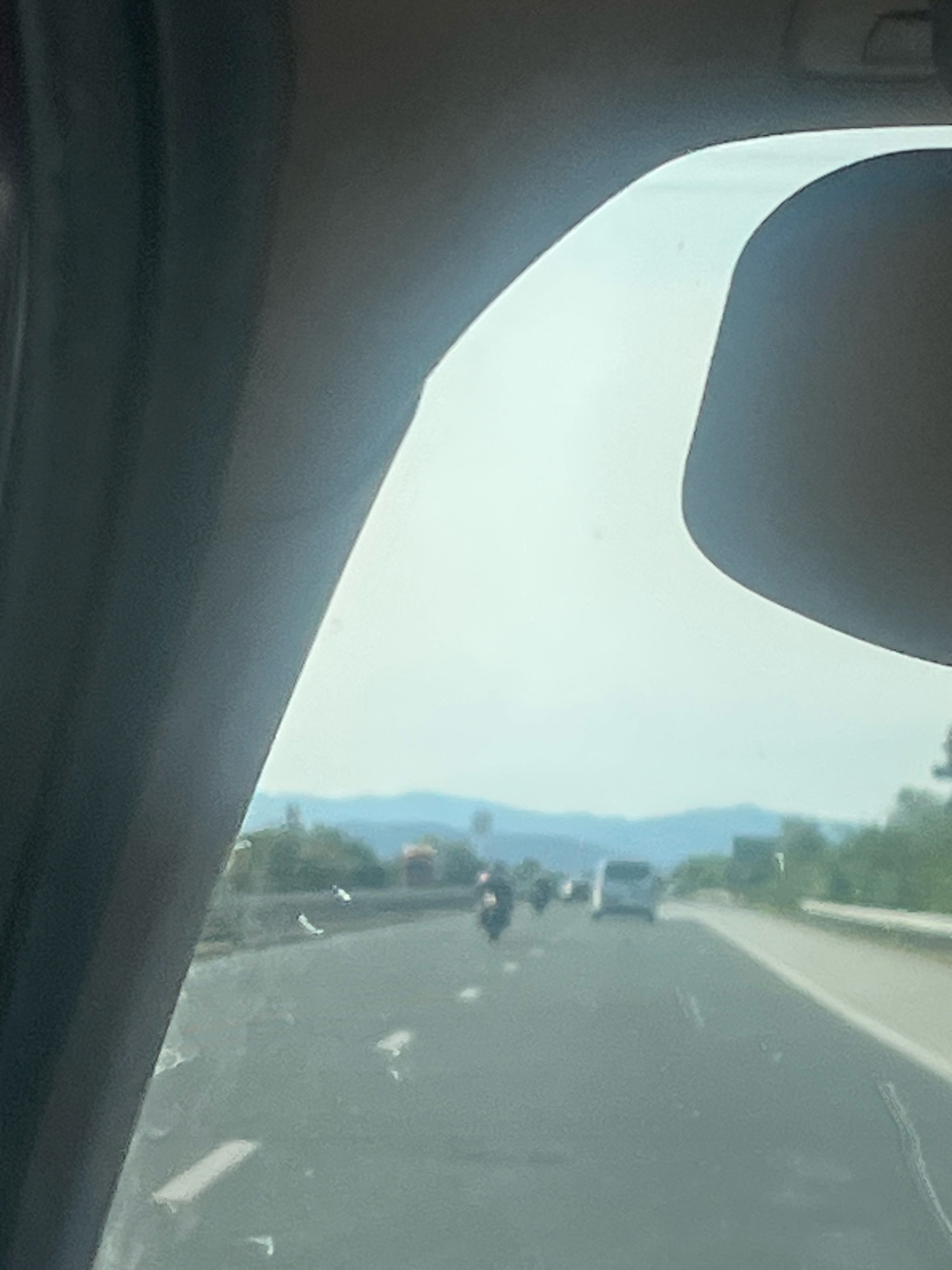 The image shows a view from inside a vehicle, looking out through the windshield. The windshield has several white streaks and smudges on it. The view outside is a highway with several vehicles, including motorcycles and cars, on it. There are mountains in the background. The lighting suggests it is daytime. The location is the city of Shtarkovo, Bulgaria.