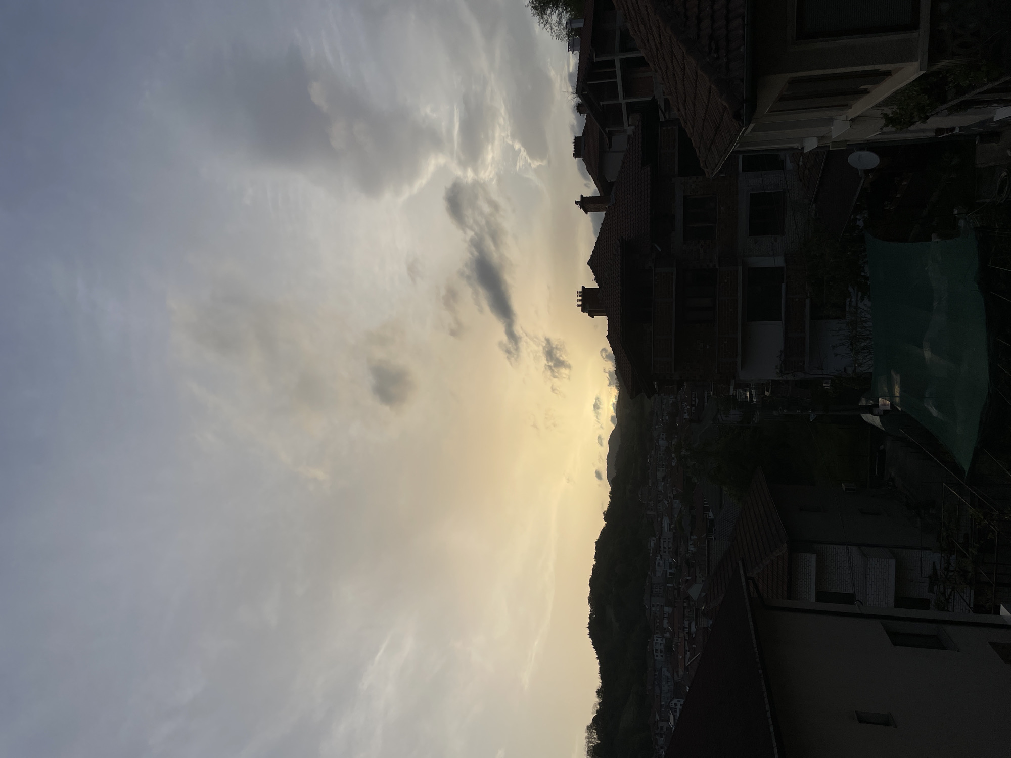 The image depicts an evening scene in Zlatograd, Bulgaria. The sky is mostly cloudy with a golden glow near the horizon. In the lower-right foreground are buildings, including a house with a red-tiled roof. Beyond the buildings, there is a hill covered with trees and houses. The silhouette of the hill separates the sky from the houses and buildings. The sky is filled with gray and white clouds.