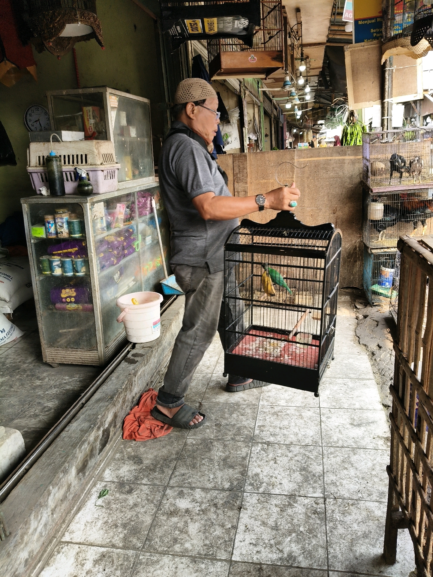 In a street market stall in Setiamekar, Indonesia, an individual wearing a grey t-shirt, grey pants, a light brown cap, and black sandals stands and holds a black bird cage containing two small birds, one green and one yellow, and two bananas. The stall features a glass display cabinet filled with various products, including canned goods and packaged items. Adjacent to the stall are multiple stacked bird cages, some containing birds. The ground is paved with grey tiles, and a red cloth is visible on the ground near the individual's feet. The overall environment appears to be an outdoor market with overhead lighting and exposed building materials.