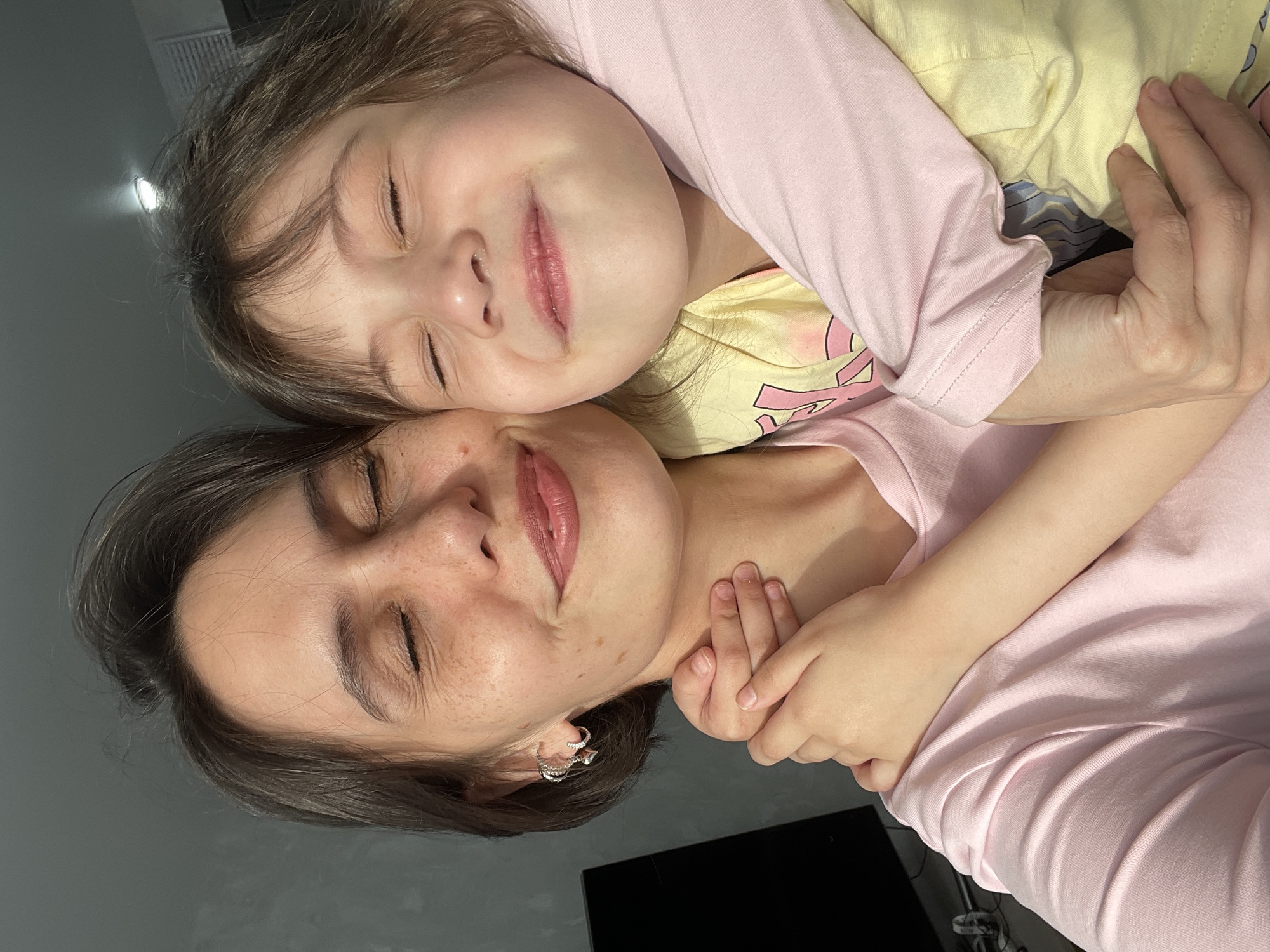 A mother and child are embracing with their eyes closed. The mother is wearing a pink t-shirt and silver hoop earrings. The child is wearing a yellow t-shirt with a pink long-sleeved shirt layered over it. The child's hand is resting on the mother's neck. The background is a plain gray wall. A corner of a black television screen is visible in the lower-right corner. It is not possible to determine if this image was taken in Izhevsk, Russia.
