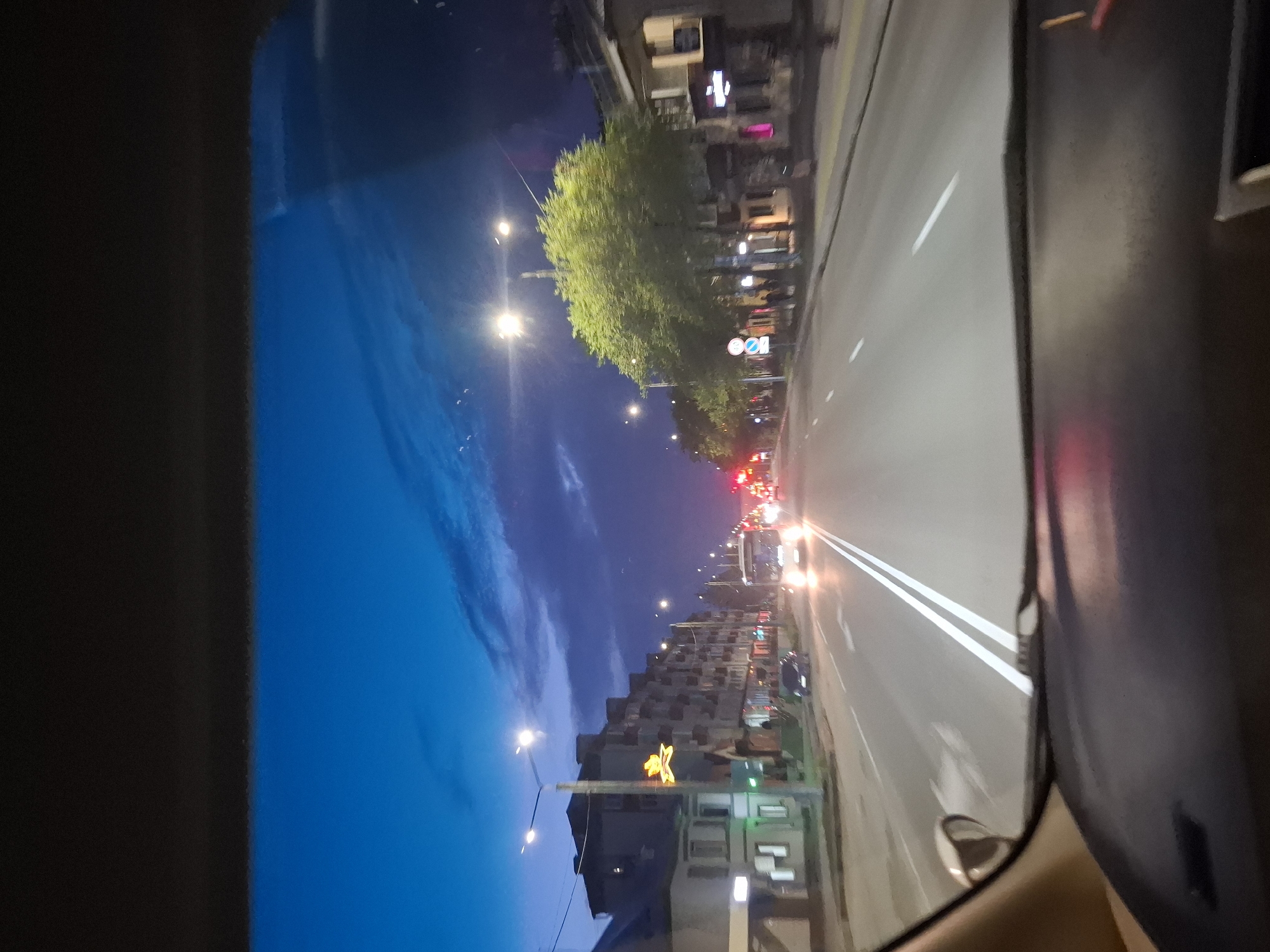 The image shows a view from inside a vehicle driving on a street at dusk or night in Svobodny, Russia. The sky is a deep blue with some wispy clouds, illuminated by artificial lights. On the left side of the street are multi-story buildings, some with lit windows and balconies. A street lamp with a yellow star-shaped decoration is visible. On the right side of the street, there are more buildings, some appearing to be shops with lit signs. Large green trees line the sidewalk. The road has multiple lanes with white lane markings, including arrows. A white bus with its headlights on is driving in the same direction as the viewer's vehicle, and other vehicles are also on the road. Traffic lights are visible in the distance. The foreground of the image shows the dashboard of the vehicle.