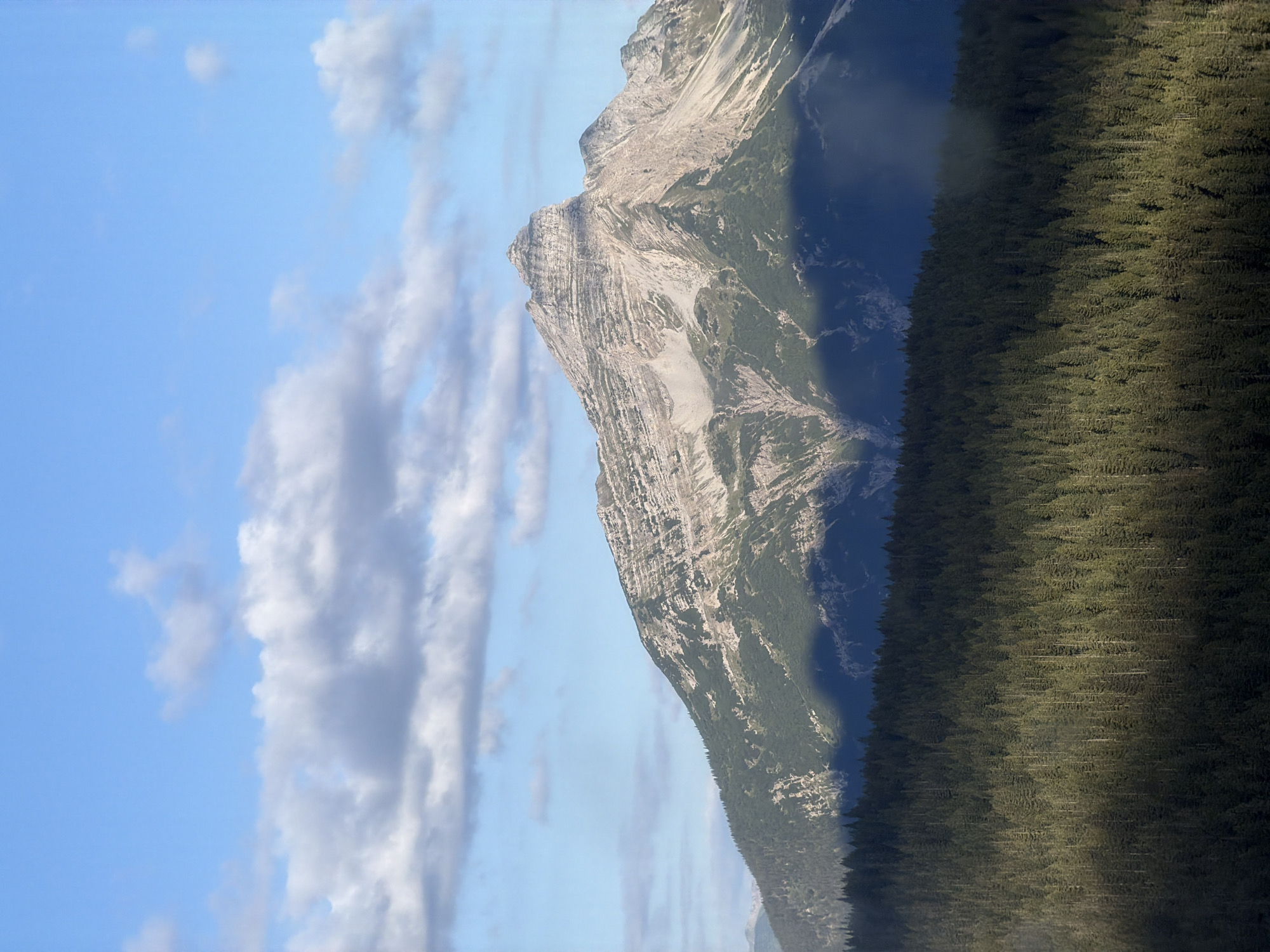 The image is a landscape photograph of a mountain range and a forest, taken from an elevated perspective. The sky is a clear blue with scattered white and grey clouds. The central subject is a prominent, rugged mountain peak with exposed rock faces showing distinct layers and striations. The lower slopes of the mountain are covered in lush green vegetation, transitioning into a dense evergreen forest in the foreground. Shadows are cast across the landscape, indicating the direction of sunlight. The image captures the natural beauty of a mountainous and forested environment. There are no people, objects, or actions depicted in the scene. The specific location in Grainau, Germany cannot be determined from the visual information alone.