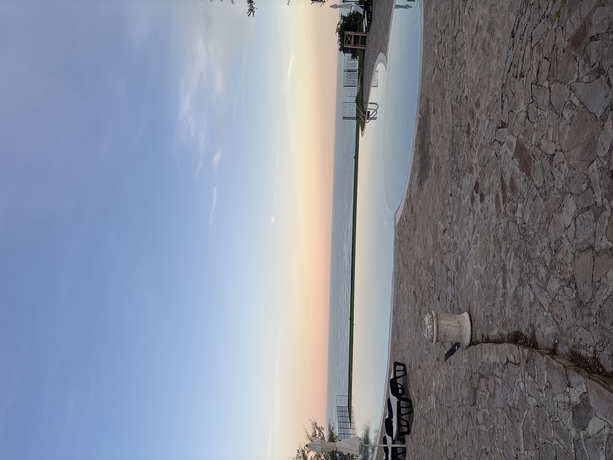 The image depicts an infinity pool overlooking the sea at sunset. The sky transitions from a clear blue at the top to soft hues of pink and orange near the horizon, where it meets the calm, reflective water. The pool's edge is lined with dark railings and further back, a fence. The foreground is paved with irregular, grey flagstones, and there are several black lounge chairs and a white umbrella. To the right, a wooden structure is visible. In the center of the foreground, a stone pedestal casts a shadow on the paving stones. The scene appears to be a tranquil resort or private property in Topola, Bulgaria, based on the provided location information. There are no people visible in the image.