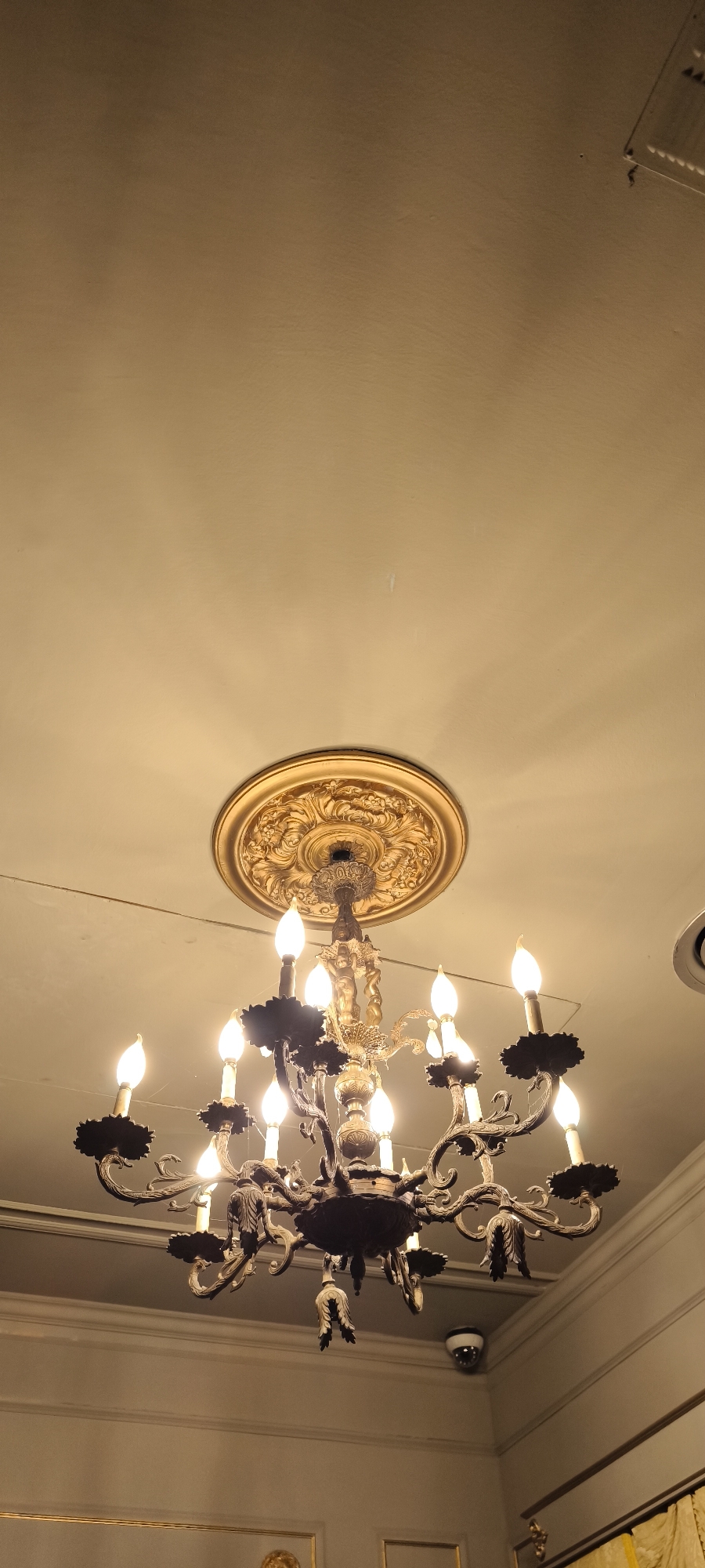 A low-angle shot shows a multi-tiered chandelier with multiple candle-shaped light bulbs hanging from a ceiling with a decorative, golden medallion. The chandelier has dark-colored ornate arms and leaf-shaped decorations. The light bulbs are lit. The ceiling appears off-white or light tan. Along the lower right edge of the image, the top of a wall with molding can be seen. A surveillance camera is visible in the background, slightly to the right of the chandelier. The location is potentially in Barcelona, Spain.