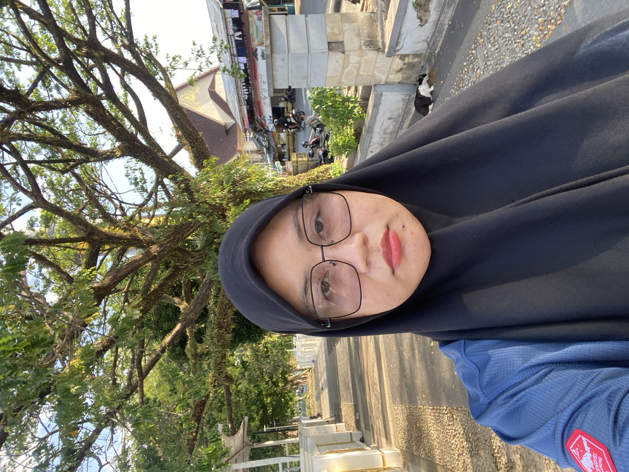 The image depicts a selfie taken outdoors in Banjarmasin, Indonesia, on a sunny day, likely in the late afternoon due to the warm lighting. The subject is a woman wearing a black hijab and glasses, and a blue collared shirt with a red and white patch on the left shoulder. She is positioned in the foreground, occupying the majority of the frame.
In the background, a large tree with extensive branches, possibly a banyan tree, dominates the upper portion of the image. Beyond the tree, a building with a reddish-brown tiled roof and decorative elements is visible. The setting appears to be a pedestrian walkway or sidewalk adjacent to a road. There are white railings and bollards lining the walkway. Several motorcycles are parked or being ridden in the background. A small black and white cat is lying on the ground in the middle ground, to the right of the image. The ground is paved with small stones and gravel. The overall environment suggests an urban area with significant green space provided by the large tree.
