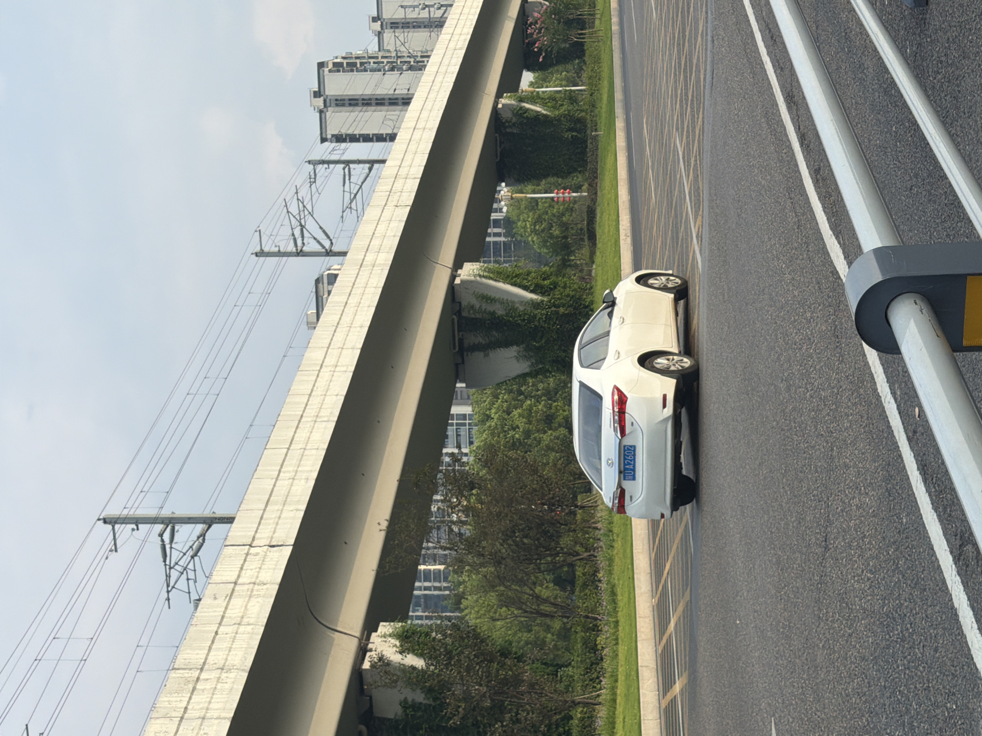 A white sedan, license plate numbered "HU A2602", is parked on an asphalt road in Jiangjin District, Chongqing, China. The car is viewed from its rear-left side. In the background, a viaduct supported by concrete pillars covered in ivy stretches across the frame. Beyond the viaduct, several tall, modern buildings are visible under a cloudy sky. A green strip of grass and trees separates the road from the viaduct's supporting structures. The scene appears to be a typical urban environment with elevated transportation infrastructure.
