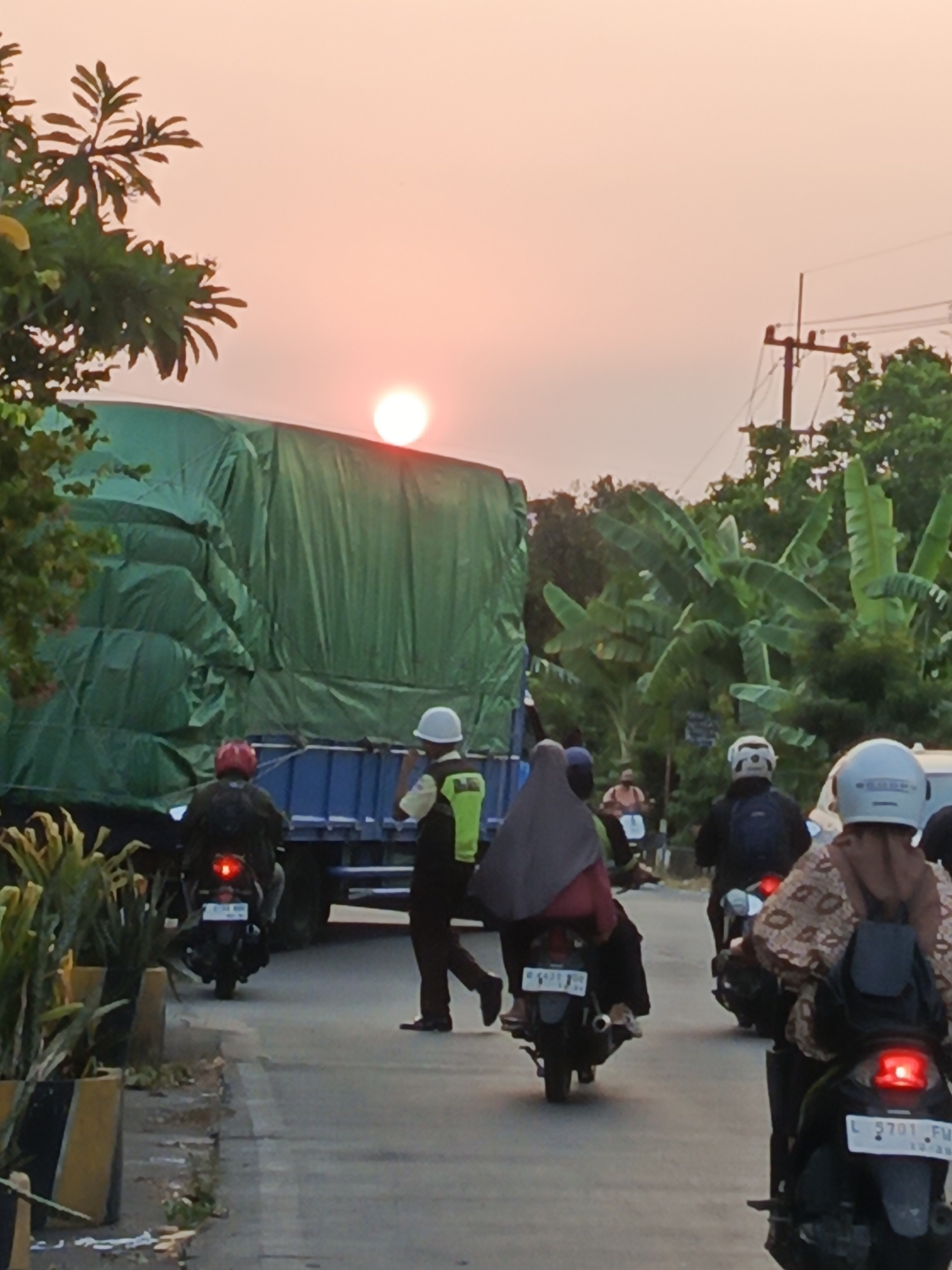 This is a vertical, medium-distance, eye-level shot of a road in Wedi, Indonesia, at sunset. The sun is visible as a bright orange orb low in the pinkish-orange sky. In the foreground, traffic is present. A large truck, covered with a green tarp, is stopped on the road. A traffic officer in a white helmet and a yellow and green vest stands next to the truck. Several motorcycles are on the road, with riders and passengers. One motorcycle in the foreground has a rider wearing a patterned brown and white top and a light blue helmet. Another motorcycle further back has a rider in a dark hijab. To the left of the truck, there are potted plants. To the right, there are trees, including banana trees, and a utility pole with wires. The overall impression is a busy street scene at dusk.