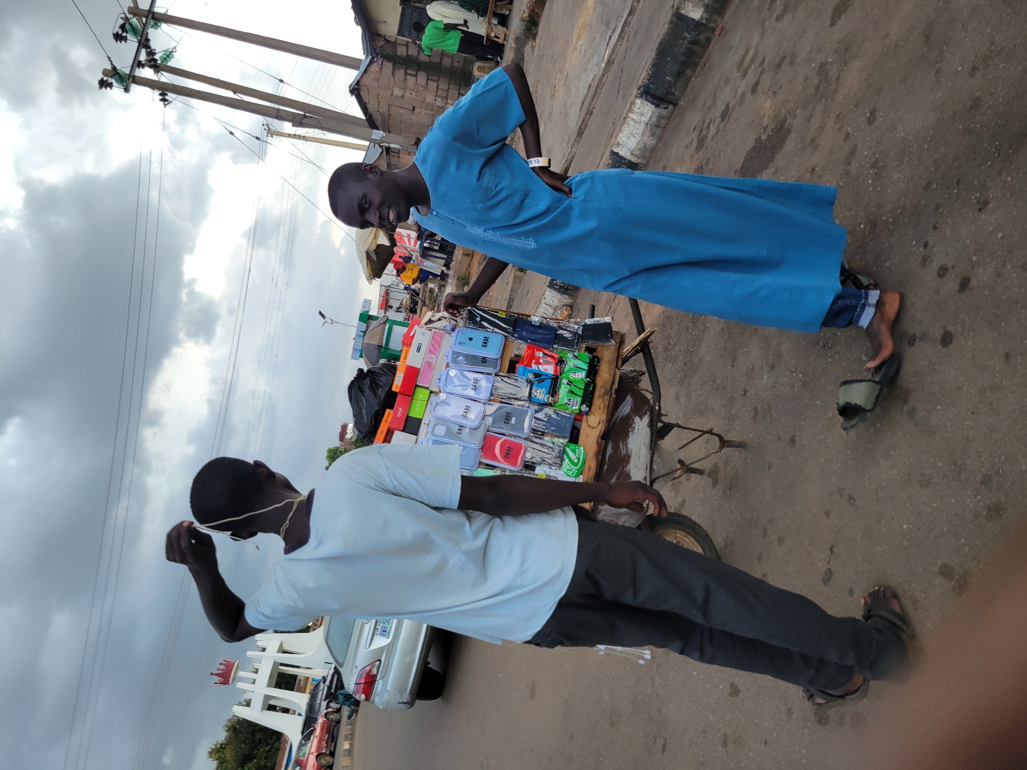 Two young men are standing on a paved street in Jos, Nigeria. One man, wearing a light blue t-shirt and dark pants, is holding a string or wire above his head, looking up at the overcast sky. The other man, dressed in a blue robe and jeans, is standing next to a cart filled with phone cases and other accessories, with his hand on his hip and smiling. The cart has a wooden frame and a metal wheel. In the background, there is a silver car, a white archway structure, and buildings. Utility poles with wires are also visible. The ground is a mix of asphalt and dirt, with some wet spots.