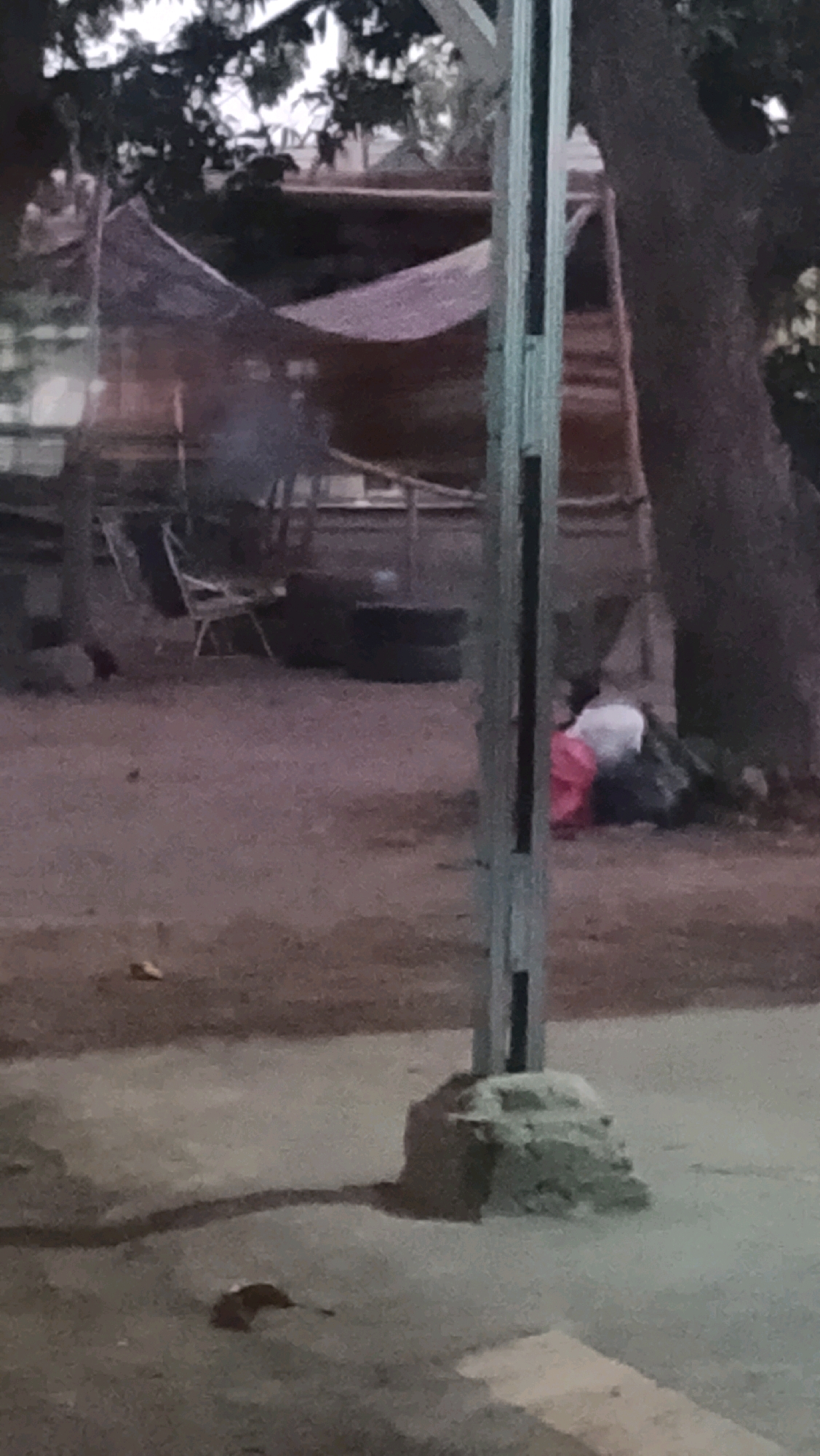The image depicts an outdoor scene at dusk, possibly in Ujungrusi, Indonesia, characterized by a rough, unpaved ground surface and ambient lighting suggesting a low-light environment. In the mid-ground, there are several figures. One individual is partially visible, lying down or seated, covered in what appears to be a white cloth, with a pink cloth beside them. They are positioned near a pile of dark, amorphous objects, possibly bags or debris. Behind this individual, a structure with a thatched or tarp-like roof is visible, supported by wooden poles. This structure appears to be some form of makeshift shelter. Adjacent to the shelter, a dark, circular object, resembling a tire, is present, with another stacked on top. Further to the left, a chair with a metal frame and a light-colored seat is visible. In the background, trees with dense foliage are prominent, and some indistinct structures are present beyond the immediate area. A vertical metal post, likely part of a street light or utility pole, stands in the foreground, with a concrete base. The overall impression is one of a modest, potentially informal settlement or roadside area.