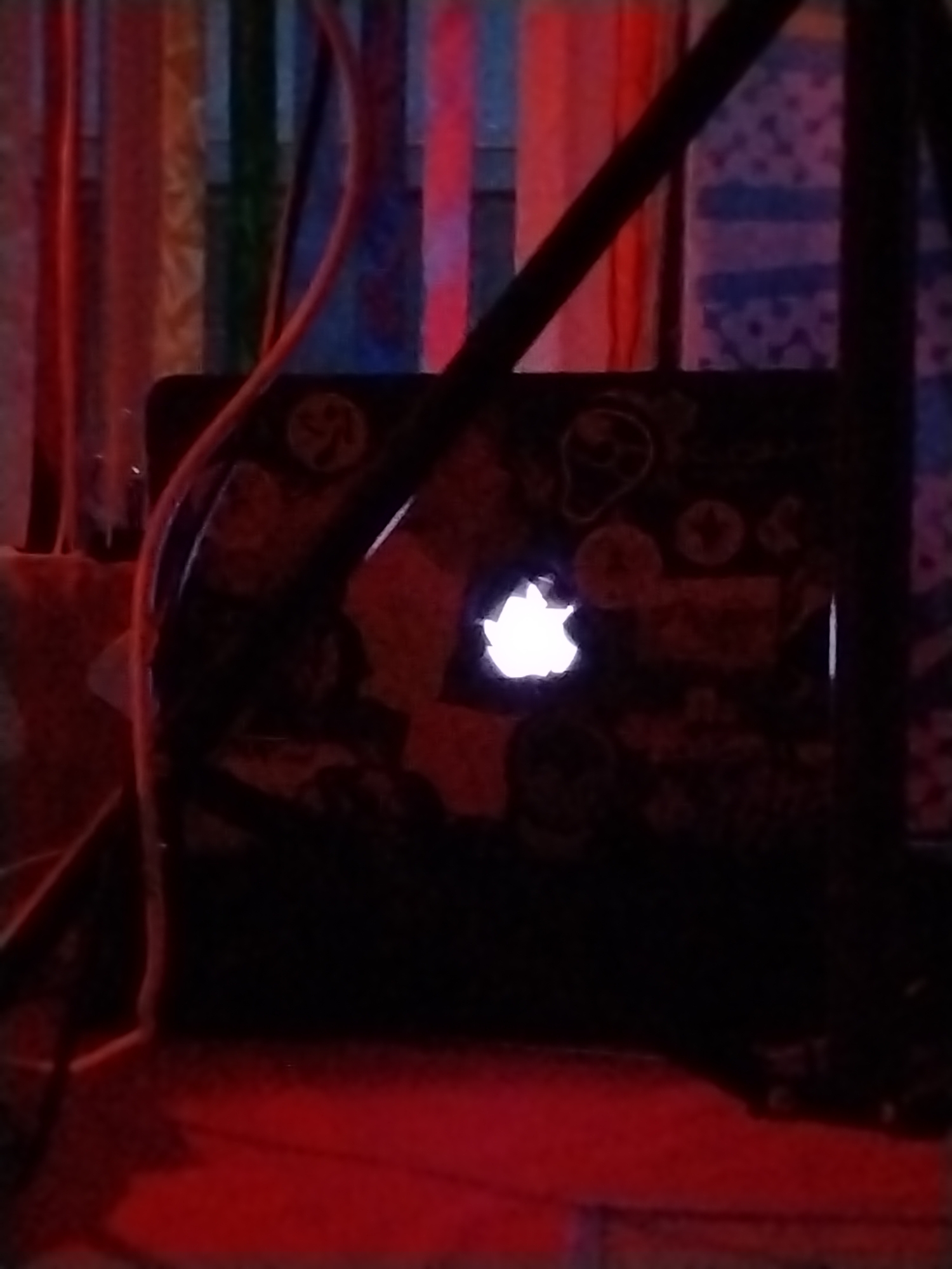A laptop computer is in a dark room. The screen is off, but the Apple logo on the lid is illuminated. The laptop is covered in stickers. The background is a blurry mix of colors and shapes. There is red light at the bottom of the scene. There is no clear indication of location in Chaikovsky, Russia, and no people are visible.