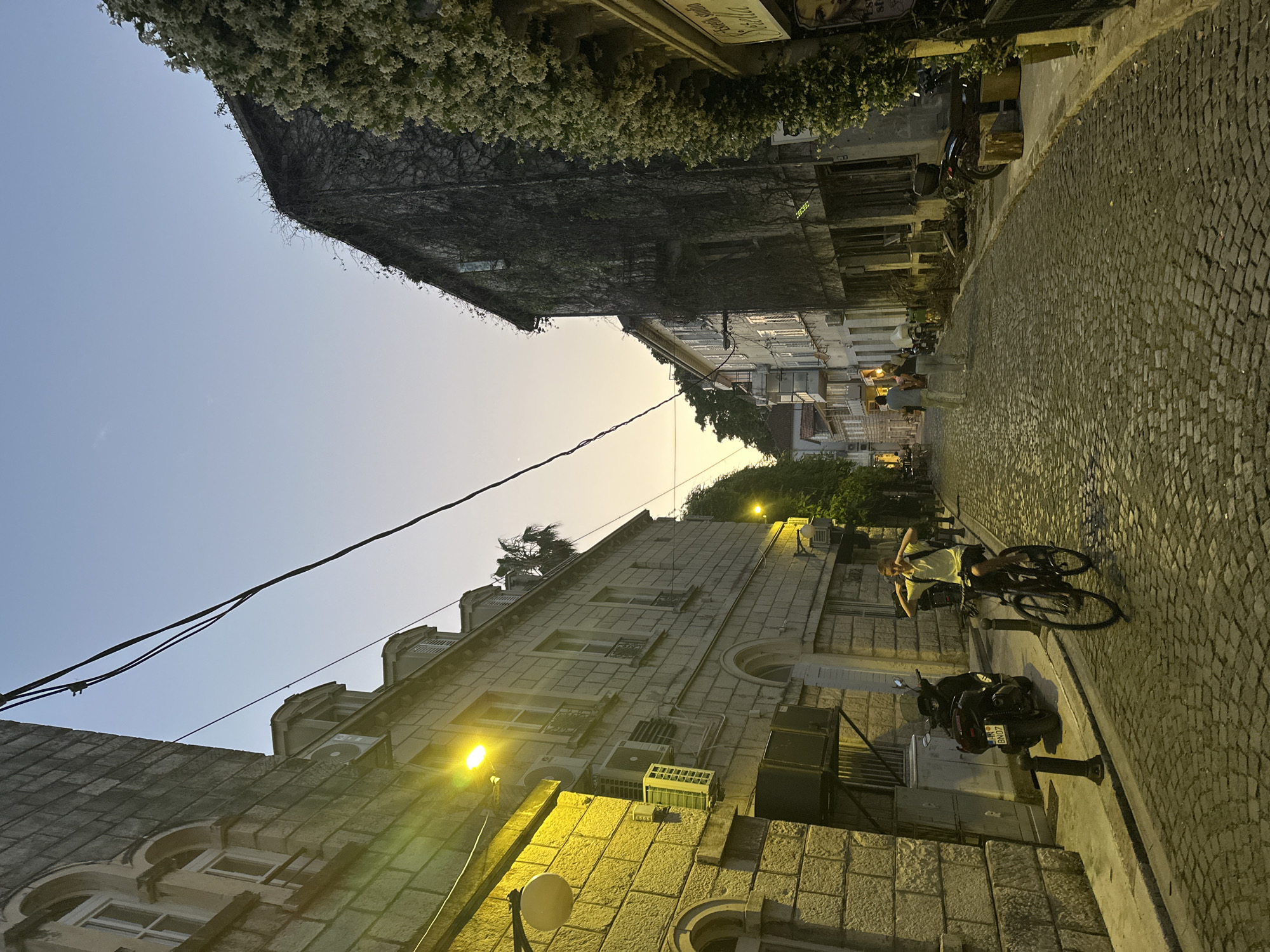 The image captures a view of a narrow, cobblestone street in Herceg Novi, Montenegro, at dusk. The sky is a pale blue transitioning to a warm orange near the horizon. Buildings line both sides of the street, one side more densely covered in vines and greenery. A person is seated on a bicycle on the right side of the street, near a parked motorbike. Street lighting illuminates the scene with a warm glow. Power lines stretch across the sky between the buildings. The street appears to be gently sloping uphill away from the viewer.