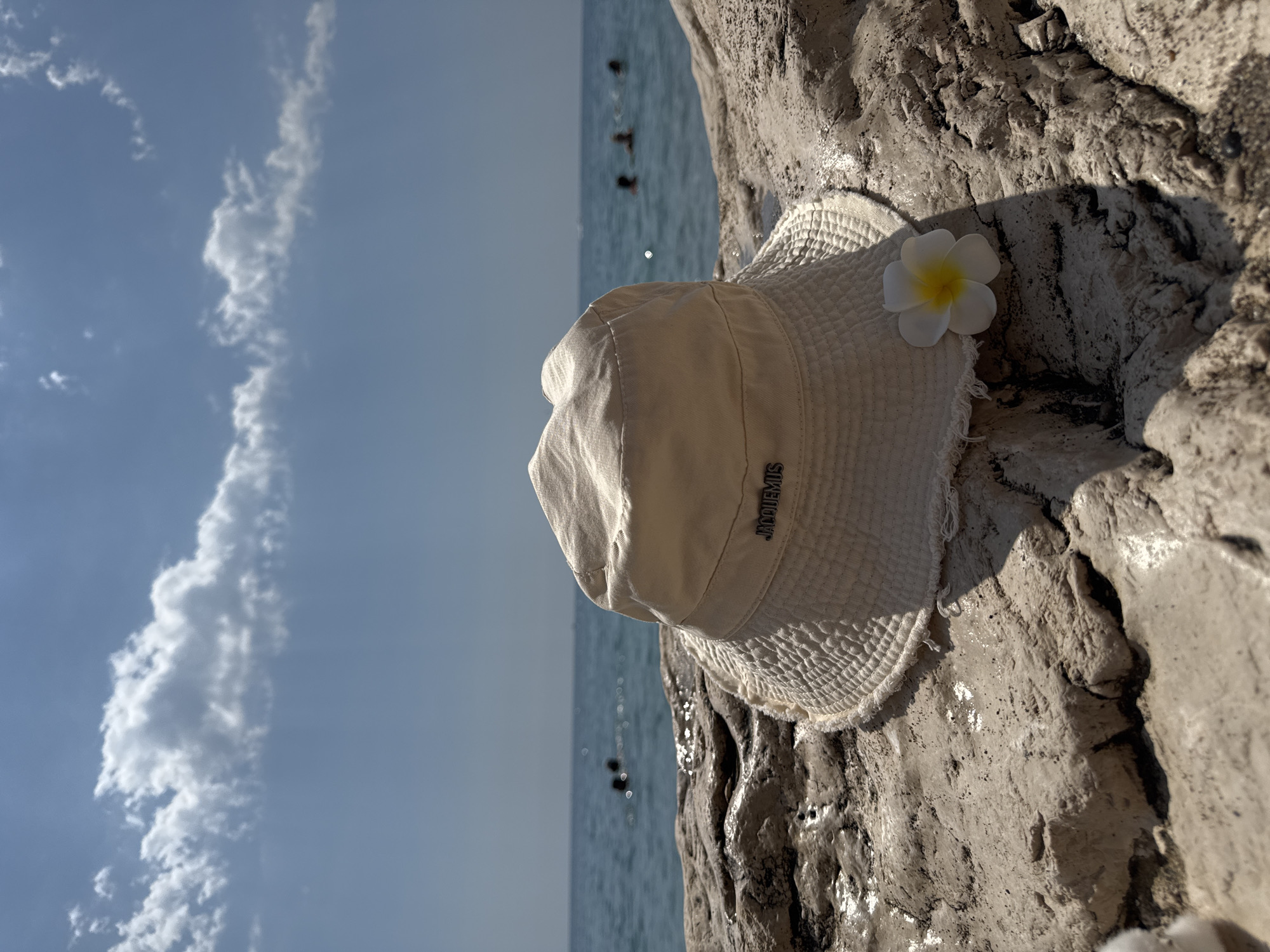 A pale beige bucket hat with a frayed edge and a small white and yellow flower rests on a textured, sun-drenched rock. The hat has a silver logo that reads "JACQUEMUS". The background features a body of calm, blue water and a sky with scattered white clouds. Several silhouetted figures are visible in the water, appearing to swim or float. The overall scene suggests a serene beach setting, likely in Kemer, Turkey, given the clear blue water and bright sunlight.