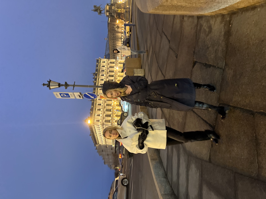 The image depicts an urban street scene at dusk or early night in Saint Petersburg, Russia, specifically in the vicinity of Palace Square. The sky is dark blue, illuminated by artificial streetlights, vehicle lights, and building illumination.
In the foreground, two blonde women are walking on a pavement composed of large, light grey rectangular paving stones. The woman on the left wears a white or light cream quilted short coat, black fitted pants, and black pointed-toe boots. She has black leather gloves on both hands and holds a small black clutch or bag with a silver chain in her left hand. Her expression is neutral, and she looks slightly to the left. The woman on the right wears a dark navy or black long trench coat with a cinched belt, a hood pulled over her head, and black knee-high boots. Her right hand is placed near her chest/collar area, and her mouth is slightly open, suggesting she might be speaking. She looks slightly to the right. Both women appear to be moving towards the viewer's right.
Behind the women, a multi-lane road carries vehicular traffic, indicated by visible headlights and taillights from several cars, including light-colored sedans and dark SUVs. A tall, dark classical lamppost stands prominently in the midground, featuring two luminaires and adorned with a blue square parking sign ("P" with "ЗОНА" and additional Cyrillic text) and a circular blue mandatory direction sign with a white arrow pointing right.
In the background, the grand, illuminated neoclassical facade of the General Staff Building, characterized by its numerous arched windows and yellowish-orange light, dominates the scene. To the far right in the background, an equestrian statue on a pedestal is partially visible. A third individual, a man in a light-colored puffer jacket and dark pants, is walking away from the camera in the mid-background, partially obscured by the lamppost. A stone wall or barrier runs along the right side of the foreground pavement.