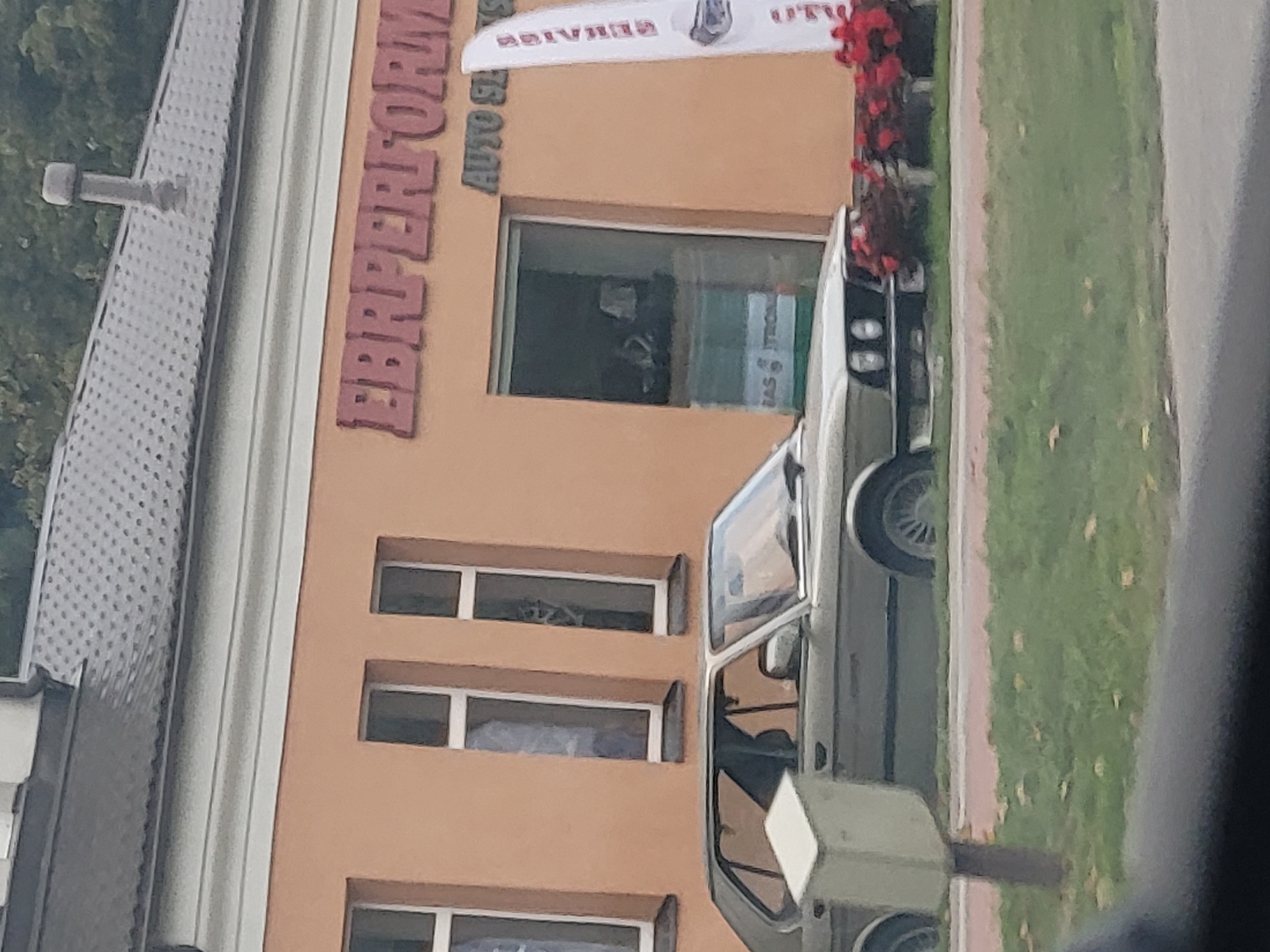 A light-colored sedan is parked in front of a building with an orange facade. The building has a dark roof and several windows. A red sign is visible on the building with text that reads "EBPerformance" and "AUTO SERVICE". A white banner with red lettering is also attached to the building. In front of the car, there is a grassy area with some fallen leaves. A light-colored mailbox is positioned to the left of the car. The car appears to be a classic model with a distinctive front grille and headlights. The overall scene suggests a commercial area, possibly an auto repair shop.