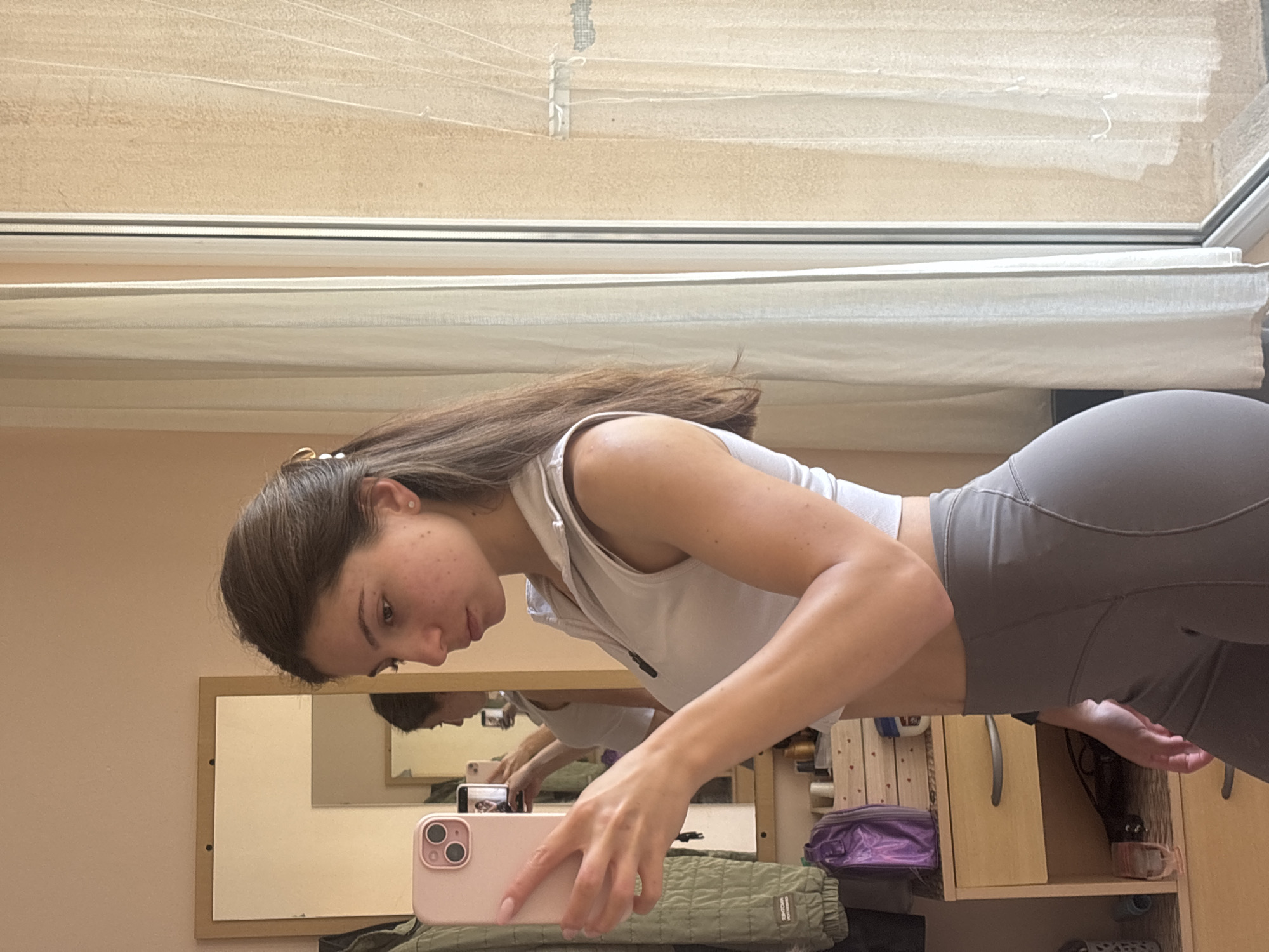 The image shows a young woman taking a mirror selfie. She is wearing a white athletic top and grey leggings. Her hair is brown and pulled back with a clip. She has freckles on her face. She is holding a pink phone with two cameras. The background appears to be an indoor setting with shelves, a mirror, and blinds on a window. The lighting suggests it might be daytime. The woman's expression is neutral, and she is focused on taking the photo. The overall impression is of a casual, everyday moment.