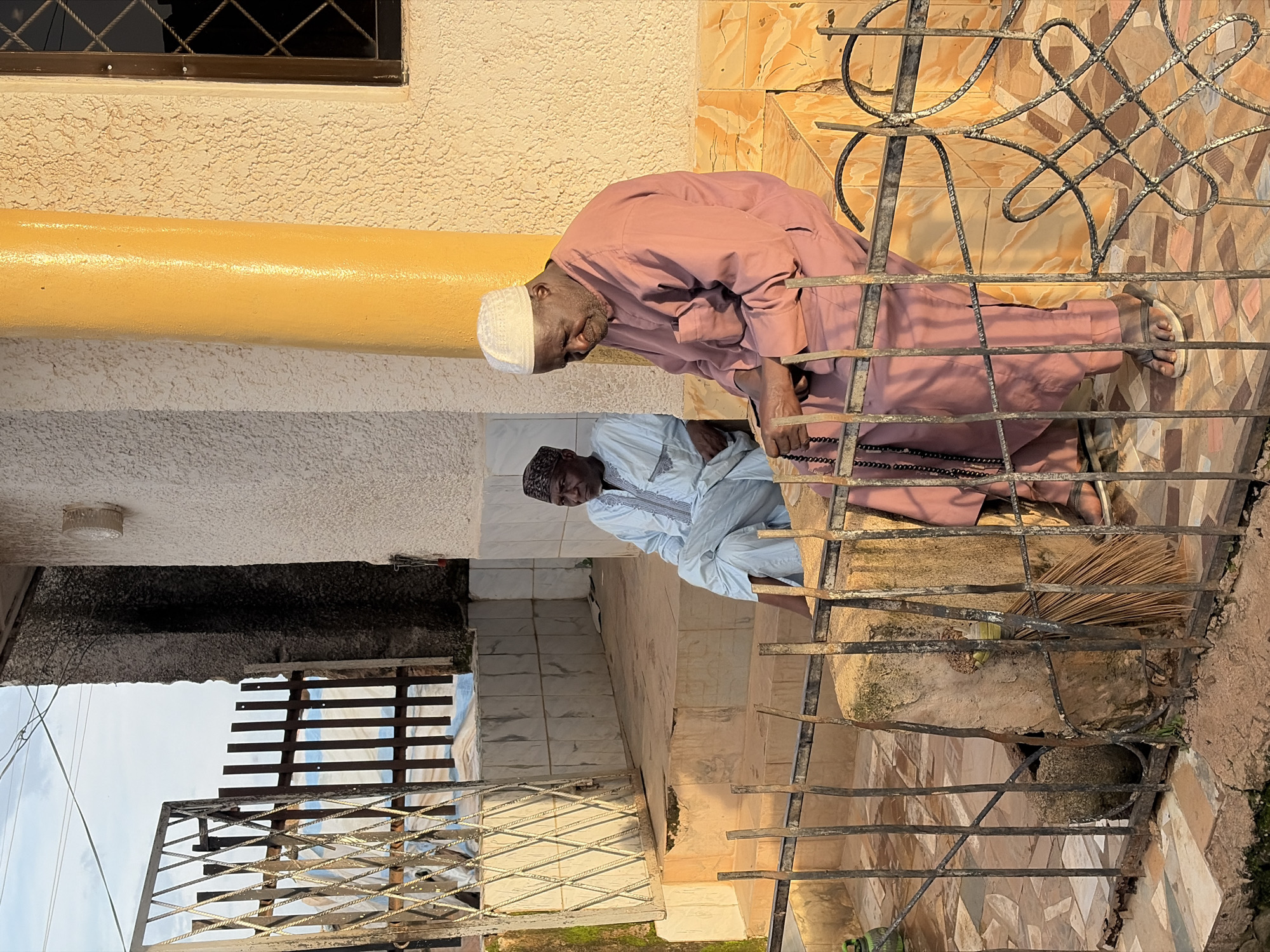Two elderly men, presumably Muslim, are seated outside a building in Jos, Nigeria. The man on the right is wearing a pink thobe and a white skullcap, holding a string of prayer beads in his hands, and appears to be in prayer or contemplation. The man on the left is wearing a light blue traditional outfit and a dark patterned skullcap. He is looking towards the right. They are seated on a tiered, tiled platform in front of a textured, light-colored wall. A bright yellow pillar is visible on the right side of the image, next to the wall. In the foreground, a black metal fence with decorative elements separates the viewer from the seated men. Behind the men, a wooden gate with metal accents is partially open, revealing a glimpse of what appears to be a pathway and some fabric. The overall scene suggests a tranquil, possibly religious, setting in an urban environment, with evidence of architectural elements and everyday objects like a broom leaning against a stone structure near the fence.