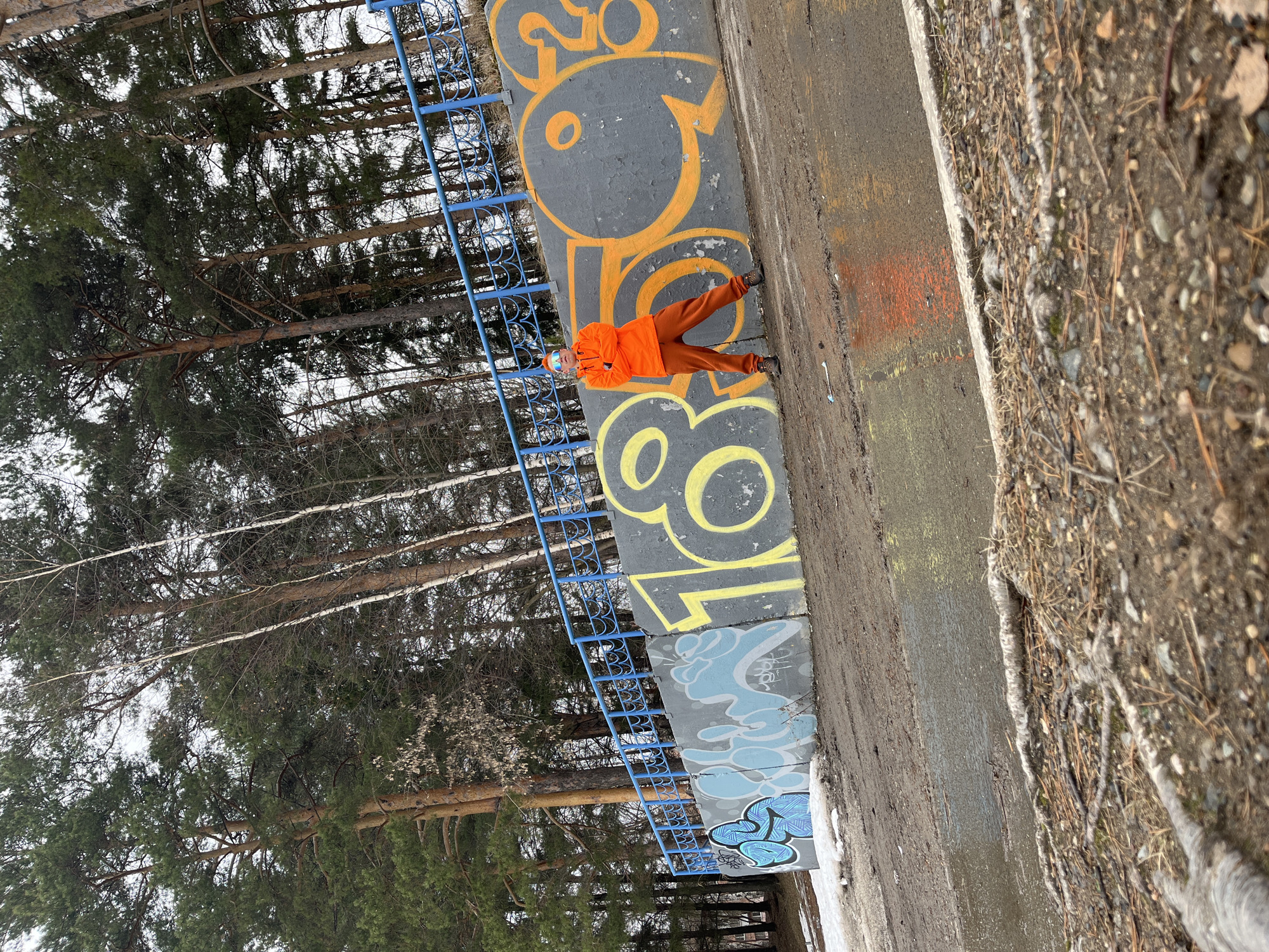 The image depicts a person dressed in an orange jacket and pants, standing next to a concrete wall covered in graffiti. The graffiti includes numbers and abstract designs. A blue metal railing runs parallel to the wall. Behind the wall and railing, there is a forest with tall trees. The ground next to the wall is covered with dirt and debris. The scene is likely located in Chaikovsky, Russia, based on the context of the prompt.