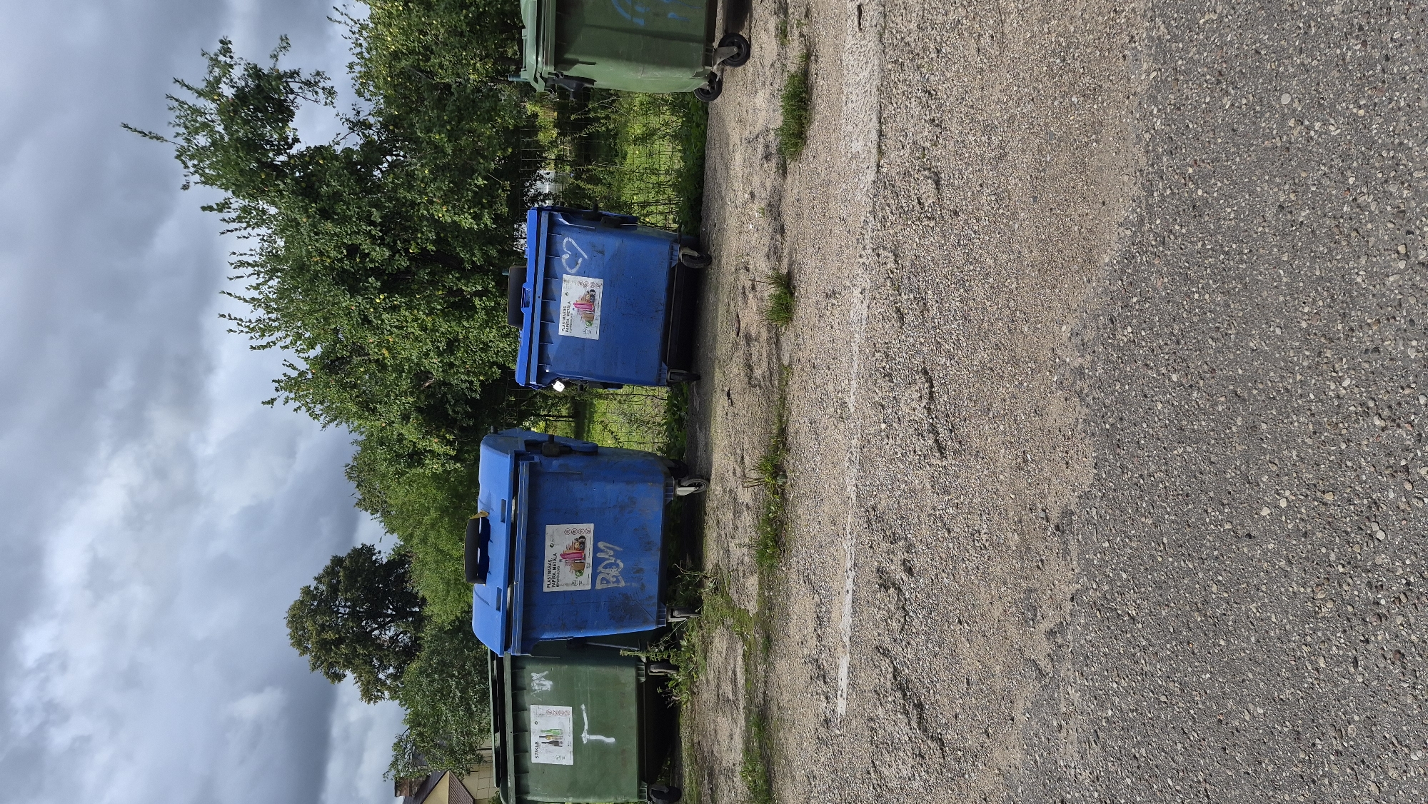 The image shows a row of large, wheeled trash bins lined up on a gravel surface. There are four visible bins: two green bins on the left and right, and two blue bins in the center. The blue bins have stickers on them, one with text and a cartoon image, and another with text and a heart symbol. The green bins also have some markings. Behind the bins, there is a low fence and lush green trees and foliage. The sky is overcast with dramatic, dark clouds. The scene appears to be an outdoor area, possibly a recycling or waste disposal point, in a semi-urban or rural setting. The general impression is one of a utilitarian space surrounded by nature under a stormy sky. This location is in the city of Krauja, Latvia.