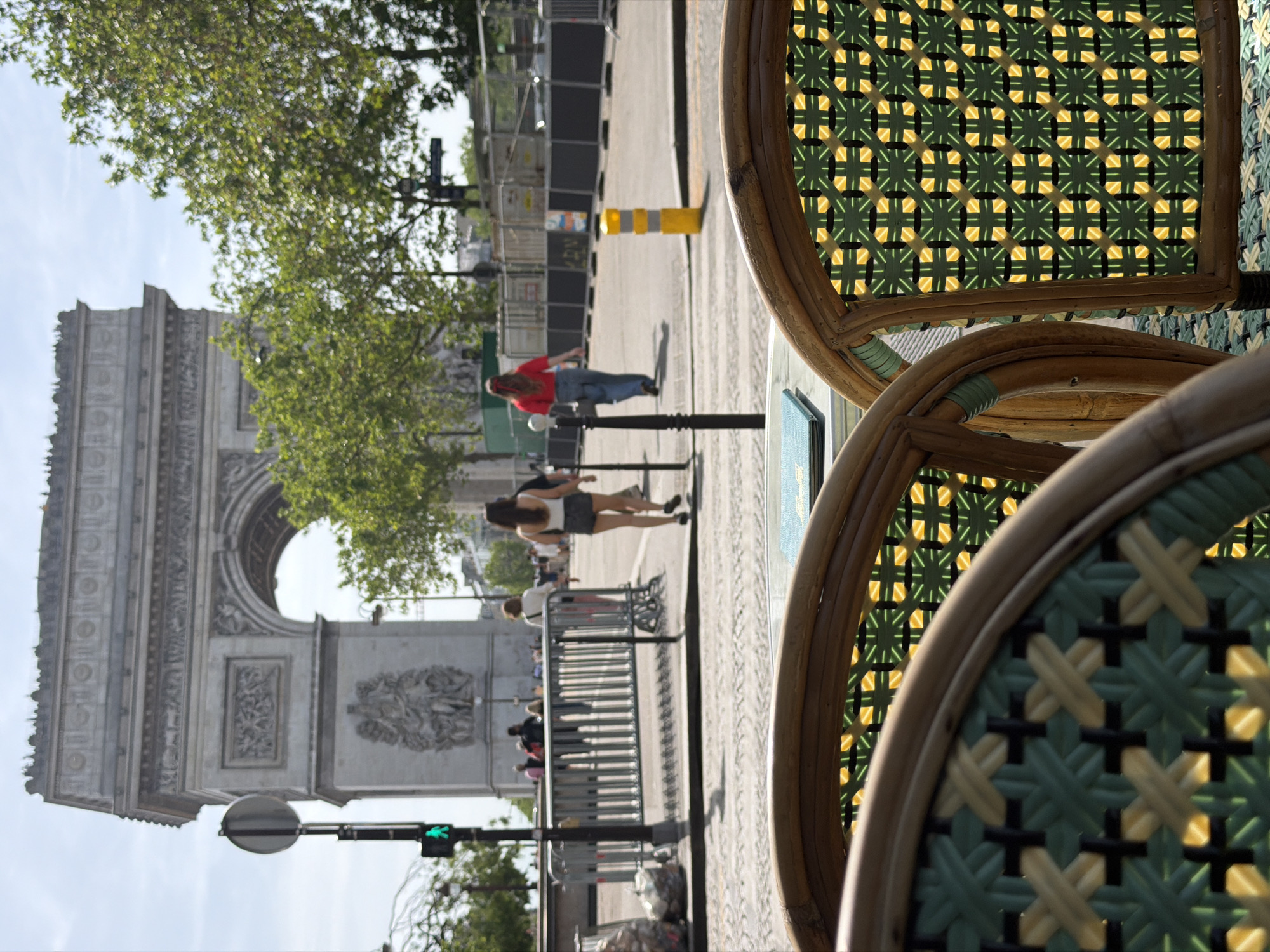 The image captures a street scene in Paris, France, dominated by the Arc de Triomphe in the background. The viewpoint is from a street-level café, indicated by the close-up view of two bistro-style chairs with woven green and beige seats. People are present on the street, including a person in a red shirt and jeans and another in a white shirt and shorts. A temporary barrier runs along the sidewalk. A yellow and white traffic marker stands in the street. A traffic signal is visible.