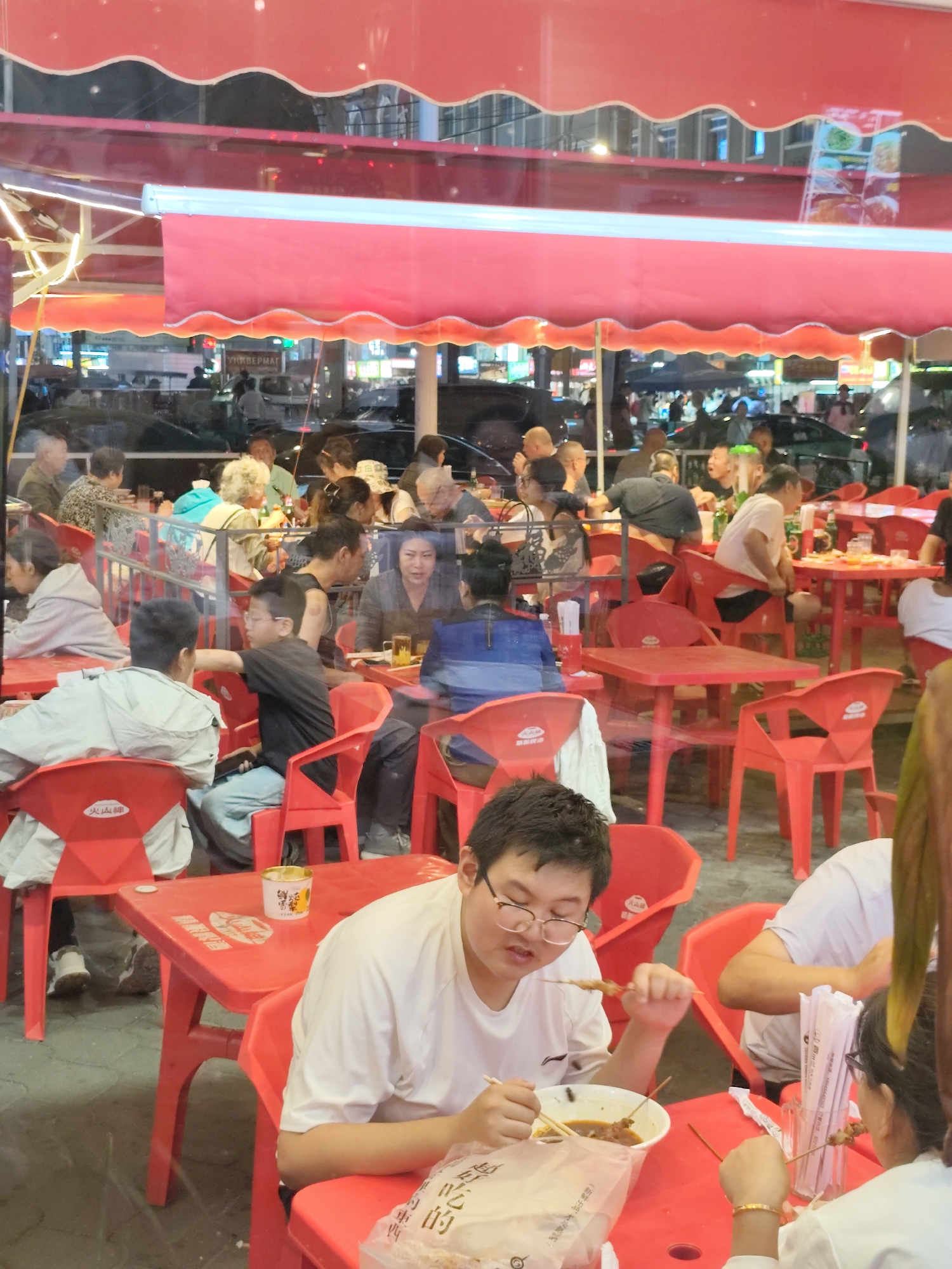 The image shows a busy outdoor dining scene at night, likely at a restaurant in Aihui, China, judging by the reflections of a sign in Chinese script on the building in the background. People are seated at red plastic tables and chairs, under red awnings. The lighting is warm and artificial, with some neon signs visible in the background.
In the foreground, a young man with glasses is eating food from a bowl with chopsticks. He is wearing a white t-shirt. To his right, a woman with dark hair is also eating. Her hand is visible, holding chopsticks, and she is wearing a bracelet. Behind them, other diners are visible, some eating and some talking. The tables are laden with food and drinks.
The overall impression is one of a lively and social gathering, with people enjoying a meal outdoors. The reflections in the glass suggest that the dining area is situated in front of a building, possibly a shop or another establishment, with traffic and pedestrian activity visible in the background. The scene is colorful and vibrant, with the red of the furniture and awnings dominating the visual palette.