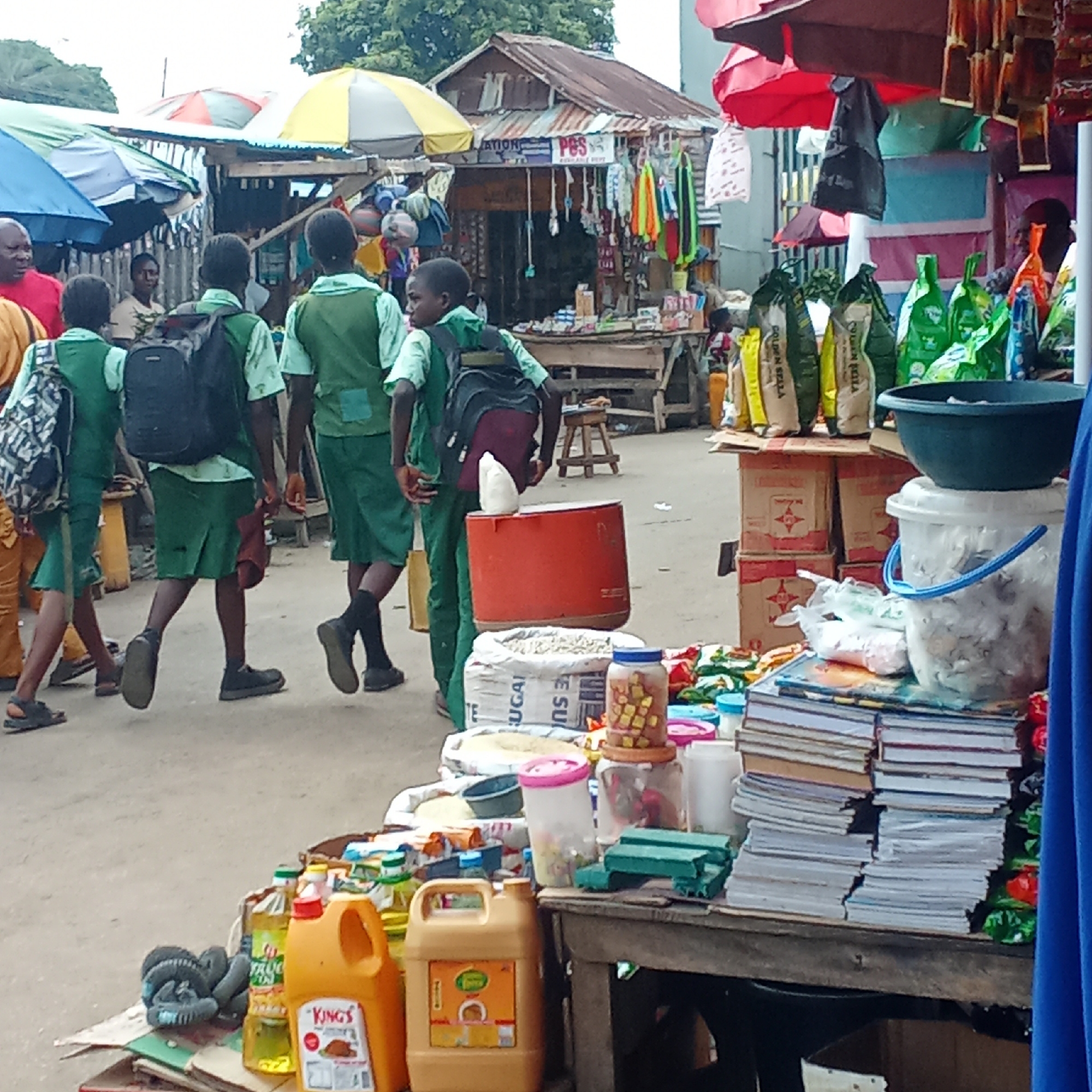 The image captures a vibrant outdoor market scene in Akwanga, Nigeria, under an overcast sky. The foreground is dominated by a market stall laden with various goods. Prominently displayed are stacks of notebooks and exercise books, jars containing small items (possibly sweets or beads), and bags of staples like rice, with visible text including "SUGAR" and "GOLDEN SELLA." Several bottles and jerry cans of cooking oil, one clearly labeled "KING'S," are also on sale. A large blue bowl rests on a white bucket, which is placed on cardboard boxes containing other packaged goods.
In the mid-ground, a group of five young students, likely on their way to or from school, navigate through the market. They are uniformly dressed in green attire—light green shirts and darker green skirts or shorts—and most carry backpacks of various colors, including red, black, and patterned designs. A large red cylindrical container is situated on the ground near them.
The background reveals more market stalls, characterized by corrugated iron roofs and colorful umbrellas providing shade. One stall exhibits a variety of hanging merchandise such as clothing and accessories, with partial text "ATION" and "PES" visible on a sign. Other adults are scattered throughout the market, some attending to stalls, others walking by, contributing to the bustling daily activity.