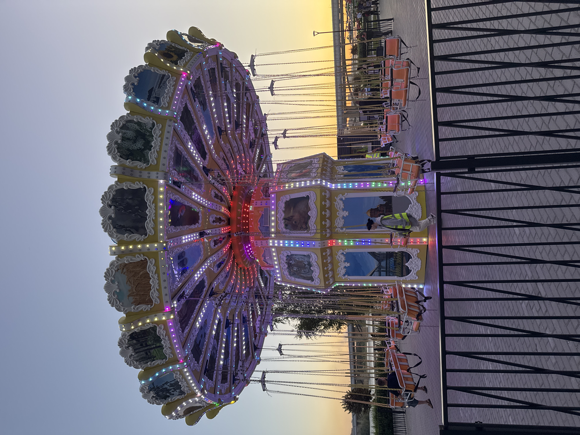 An amusement park swing ride is illuminated with colorful lights against a dusk sky in Novosibirsk, Russia. The ride features a circular canopy decorated with images and ornate trim, from which numerous chains suspend orange seats. Several people are seated on the ride, and a few others are walking around the perimeter of the ride. The ground is paved with interlocking bricks, and a black metal fence separates the viewer from the ride. In the background, a body of water and a bridge can be seen, along with the lights of the city. The overall impression is one of a lively evening at an amusement park.