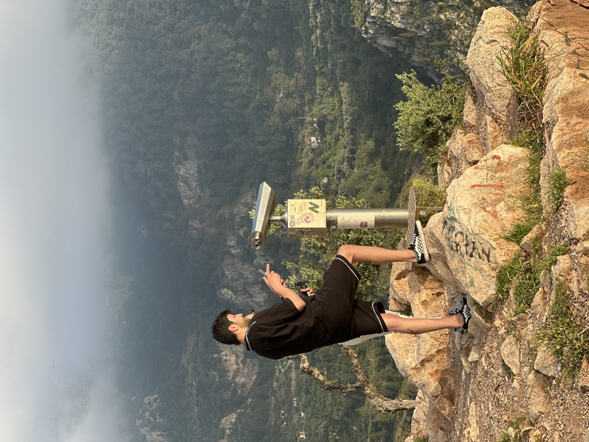The image depicts a young man standing on a rocky cliffside, gazing out at a vast, mountainous landscape in Fethiye, Turkey. The man, with dark hair and a beard, is dressed in a black short-sleeved shirt with white trim, black shorts with white piping, and black and white checkered Vans shoes. He is positioned next to a coin-operated binocular viewer, his right hand raised with an index finger pointing, as if indicating a point of interest in the distance. He appears to be holding a smartphone in his left hand.
The setting is a high vantage point, characterized by rugged, light-colored rocks and scattered green vegetation, including small bushes and wildflowers. Some rocks show signs of graffiti, with partial words like "GRYAN" visible. The binocular viewer's stand has several stickers, including a distinct yellow one featuring a white 'M'.
Beyond the immediate foreground, a sprawling valley unfolds, densely covered with green forest. A cable car system is visible in the distance, with a white gondola suspended above the trees. The left side of the frame is enveloped in a soft, white mist or low clouds, partially obscuring the distant mountains and creating a serene, hazy atmosphere. The scene is illuminated by daylight, suggesting a clear but potentially foggy or hazy day. The overall impression is one of breathtaking natural beauty and a popular tourist viewpoint.