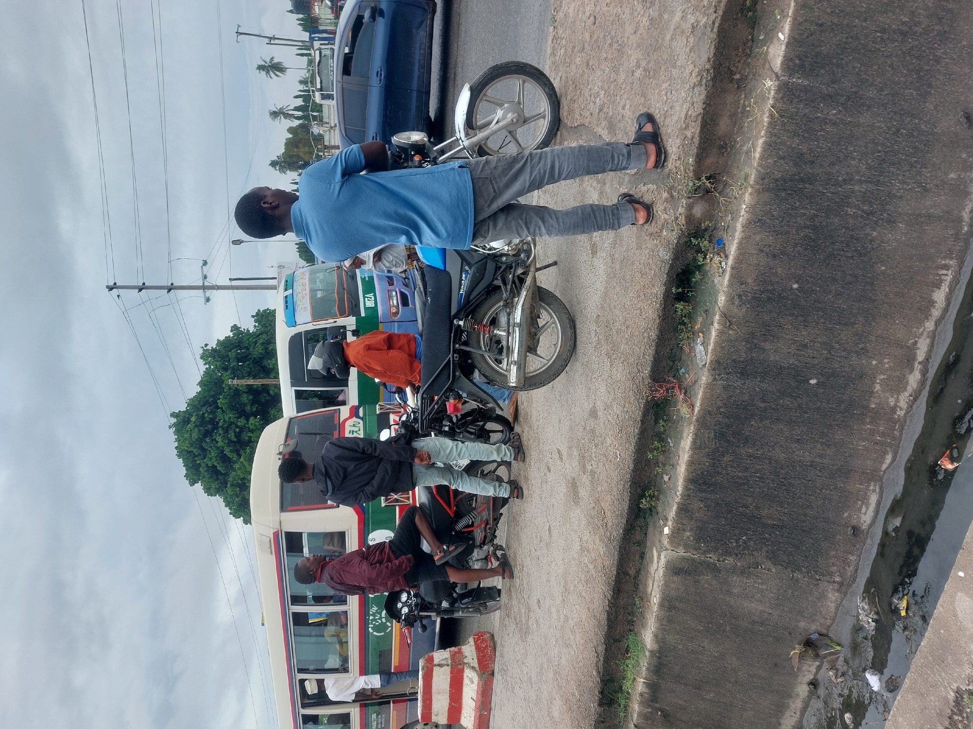 The image shows a street scene in Dar es-Salaam, Tanzania. Several men are standing near motorbikes along the side of a road. Behind them are buses and a blue car. Overhead, there are power lines. The edge of the road is concrete, and a drain runs alongside it with visible debris. The sky is overcast.