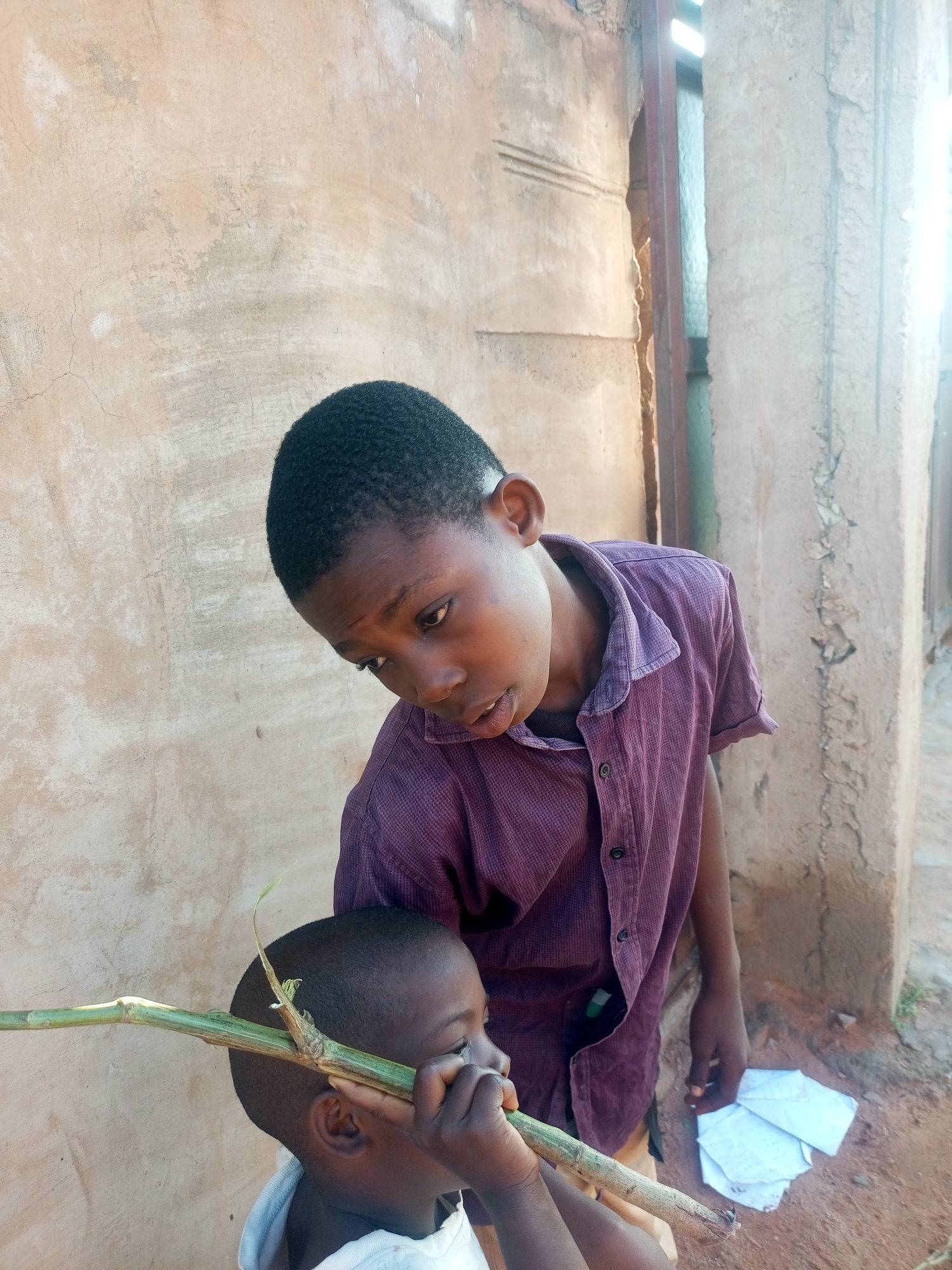 In a dusty alleyway with a rough, concrete wall as the backdrop, two young Nigerian boys are engaged in an interaction. The elder boy, wearing a purple, short-sleeved button-down shirt, leans in, his gaze directed downwards at the younger child. His expression is one of close attention or possibly explanation. The younger boy, facing away from the camera and partially obscured, holds a long, green stalk horizontally across his face, as if he were playing a game or pretending to smoke it. He wears a light-colored garment, possibly a shirt. In the background, a narrow opening reveals a glimpse of a metal frame and more of the textured wall. A few crumpled white papers lie on the dusty ground to the right of the elder boy. The scene suggests a moment of childhood play or shared activity in a simple, outdoor urban environment.