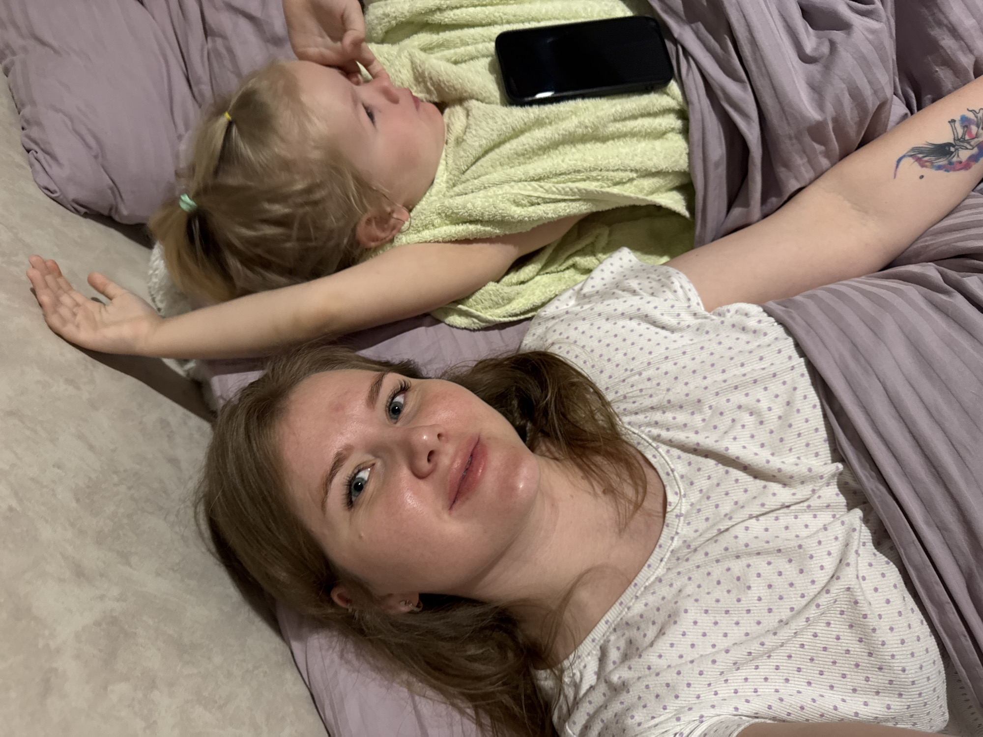 An adult woman and a young child are lying on a bed or sofa in an indoor setting, presumed to be in Gornyi (Dvurechenskoe S/P), Russia. The woman, with light brown hair and blue eyes, is in the foreground, looking directly at the camera with a relaxed expression and a slight smile. She is wearing a light-colored top with purple polka dots and horizontal stripes. A colorful tattoo is visible on her left forearm.
Behind her, a young child with blonde hair, styled in two small ponytails with yellow and light blue elastic bands, is lying on their back. The child is covered with a light green terry cloth towel or blanket and appears to be looking upwards and slightly away from the camera. A black smartphone is placed on the green towel near the child's head.
The bedding consists of muted purple or lavender fabric, some sections appearing striped. To the left, a lighter, possibly beige or off-white textured surface is visible, which could be part of the furniture or wall. The lighting is soft, suggesting an indoor environment, possibly in the evening or a dimly lit room. The overall atmosphere conveys a sense of calm and relaxation, possibly a shared quiet moment between the two individuals. There is no visible text in the image.