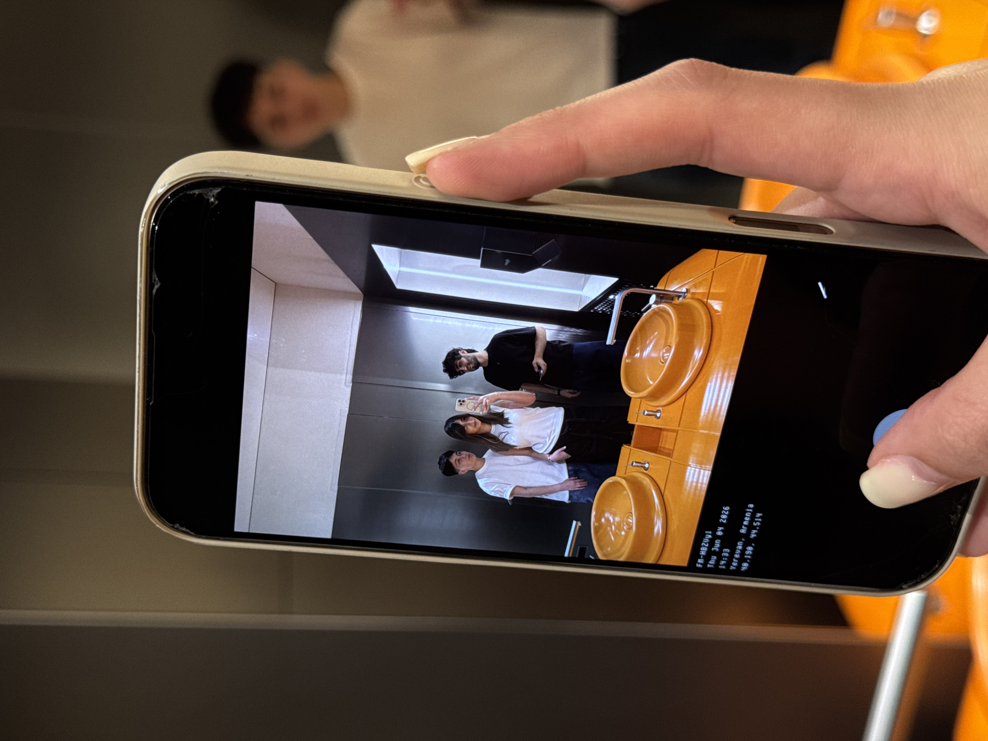 A close-up shot shows a hand holding a smartphone horizontally, capturing the image displayed on its screen. The phone has a light-colored case, and the edges of the screen protector show some wear or small cracks. In the background, out of focus, is a person wearing a white shirt.
The smartphone screen displays a photograph of three individuals inside what appears to be an elevator or a room with reflective walls. From left to right, there are two men and one woman. The woman, positioned in the middle, is holding a smartphone up to take a selfie of the group. The man on the far right has dark, curly hair and a beard, wearing a black shirt and dark pants. The woman wears a white t-shirt and dark pants. The man on the far left wears a white t-shirt and blue jeans. To the right of the people on the screen are two distinctive yellow, bowl-shaped structures, possibly decorative sinks or basins, set into a matching yellow counter or furniture. The scene suggests a casual and social moment.
A portion of the smartphone's camera interface is visible on the screen, including a blue-tinted shutter button on the bottom right. An informational text overlay is present on the lower left of the screen, which reads: "FM-HBZ0y1," "14:33," "Thu Jun 04 2026," "Yerevan, Armenia," and "40.198, 44.514." This indicates the photo on the screen was taken on June 4, 2026, at 2:33 PM, in Yerevan, Armenia, with the specified geographical coordinates.