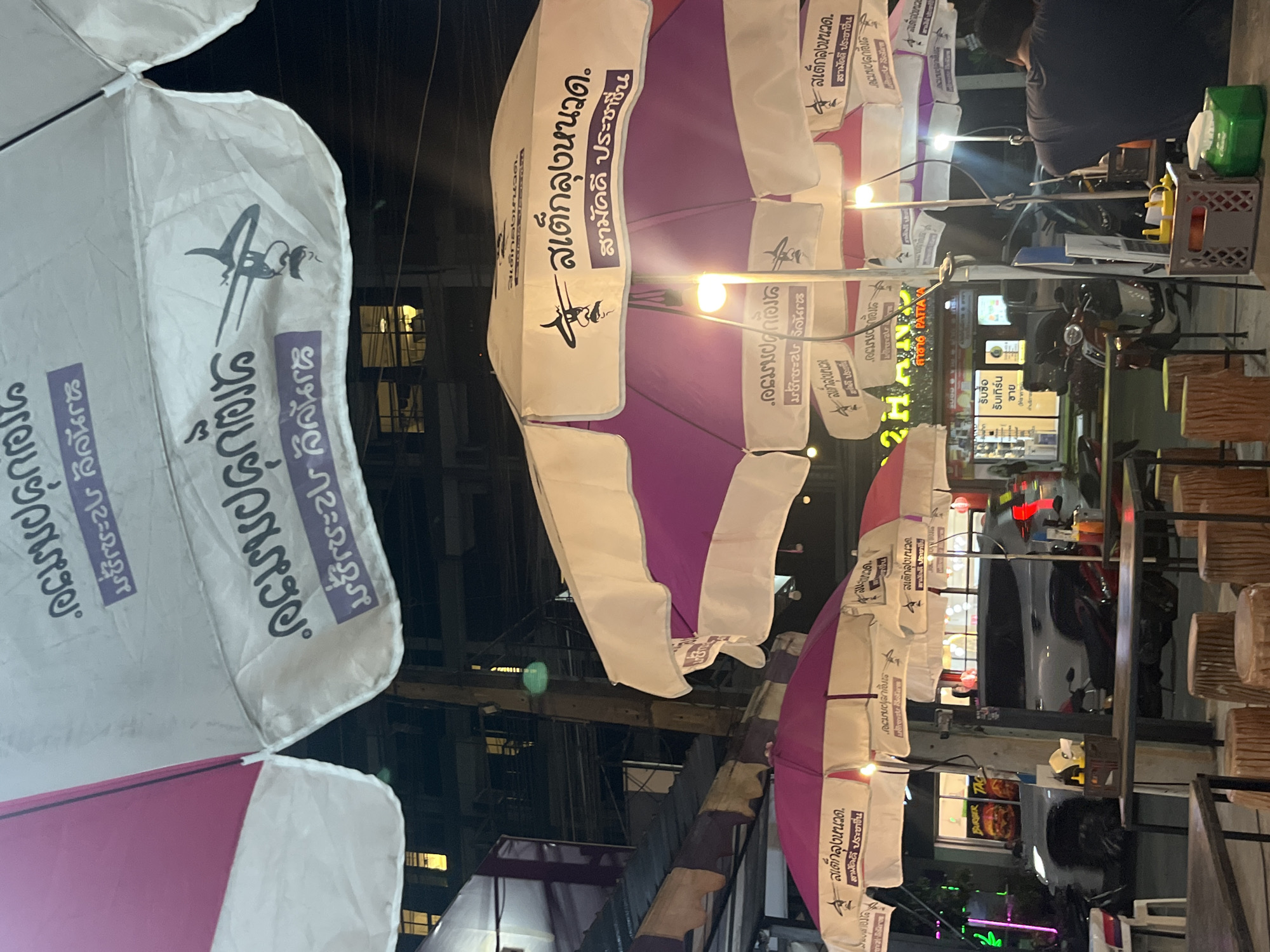 The image shows a street scene at night in Pattaya City, Thailand. Several pink and white umbrellas, printed with Thai script and the words "สเต็กลุงหนวด สามัคคี ประชาชน", are visible. Street lighting casts a glow on the scene. Behind the umbrellas, there are buildings, vehicles, and a person standing at a food stall. There are signs for "Burger" and "16 PATTAYA." The area appears to be a street food vendor setting.