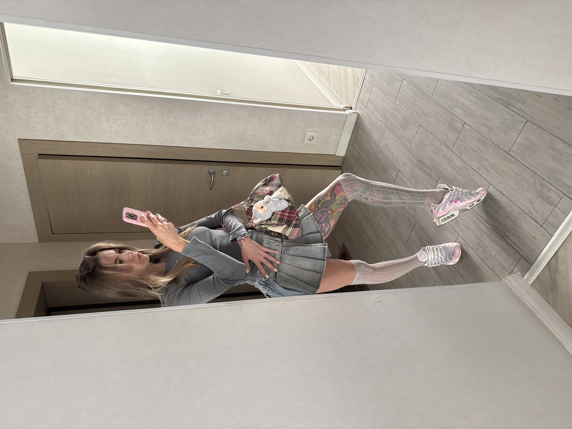 A woman is standing in a hallway and taking a mirror selfie with her pink smartphone. She is wearing a grey long-sleeved shirt, a denim pleated skirt, white thigh-high socks, and pink and grey sneakers. She has tattoos on her legs and is carrying a plaid handbag with a stuffed toy attached to it. The hallway has light grey wood-plank flooring and beige walls, with two closed doors visible. The location is identified as Moscow, Russia.