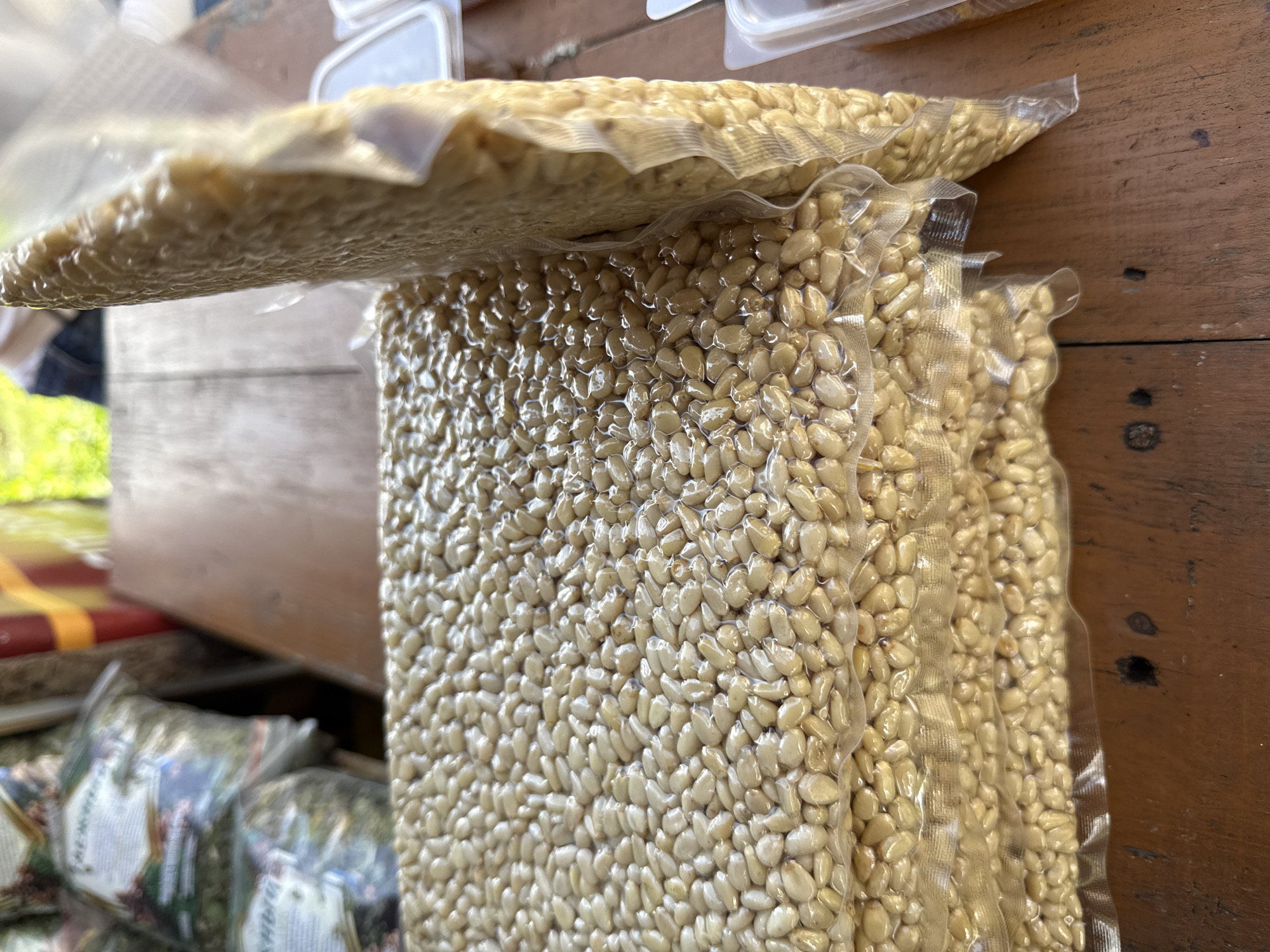 In Chemal, Russia, a close-up shot focuses on several vacuum-sealed packages of pine nuts stacked on a wooden surface. The packages are clear, allowing the light-colored, elongated pine nuts to be fully visible. The surface is a weathered wooden table with visible grain and occasional dark marks. In the background, out of focus, are more packages of dried goods, some with labels featuring text and images of nature. There are also hints of greenery and people in the far background. The lighting suggests it is daytime, possibly outdoors or in a well-lit indoor market.