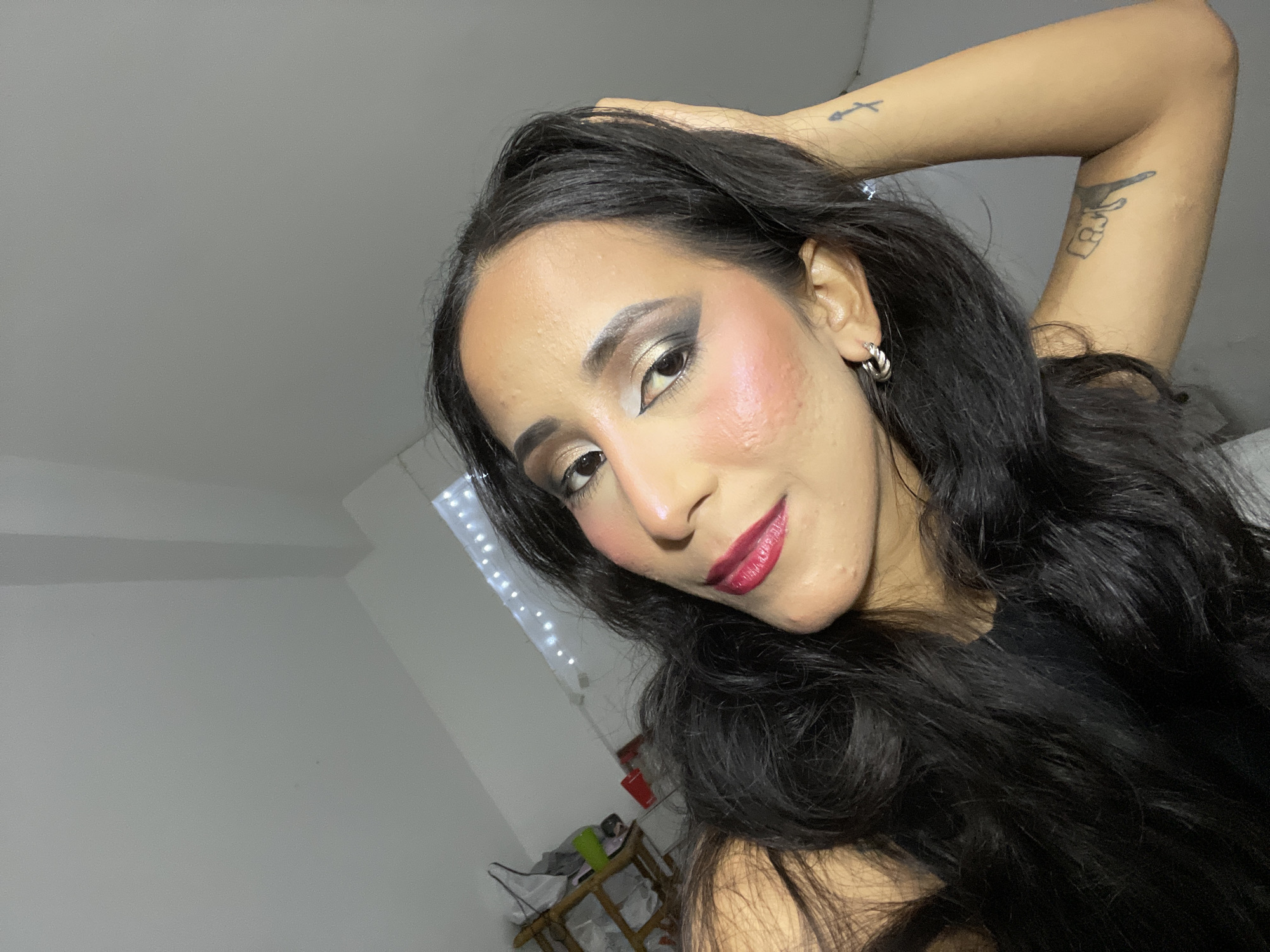 In a dimly lit interior room, a woman with long, dark hair is captured in a close-up, selfie-style photograph. She is wearing makeup, including dark eyeshadow, eyeliner, and bold red lipstick. Her right arm is raised, with her hand resting on the back of her head. There are two tattoos visible on her right arm: one resembling an arrow and the other a hand with a number. She is wearing a silver hoop earring in her visible ear. The background appears to be a plain wall, with a window to the left that has string lights. A shelf with some objects is partially visible in the lower left quadrant of the image. The overall lighting suggests the photograph was taken indoors, possibly at night or in a room with limited natural light. The location of Porlamar, Venezuela is not discernible from the visual information provided in the image.