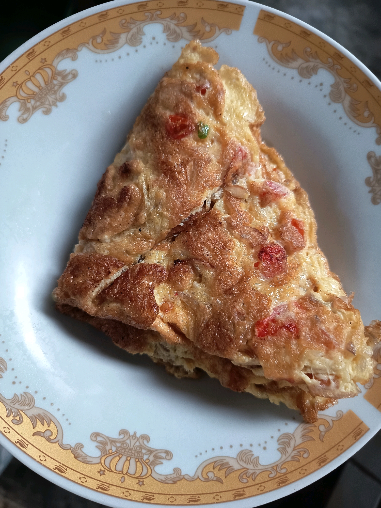 A close-up shot shows a folded omelet on a white plate with gold trim. The omelet is golden brown with visible pieces of red tomato and green chili pepper. The plate has an ornate gold pattern around its rim, featuring scrollwork and crown-like motifs. The background is blurred and dark, suggesting an indoor setting. This image was captured in Aluekumba, Indonesia.