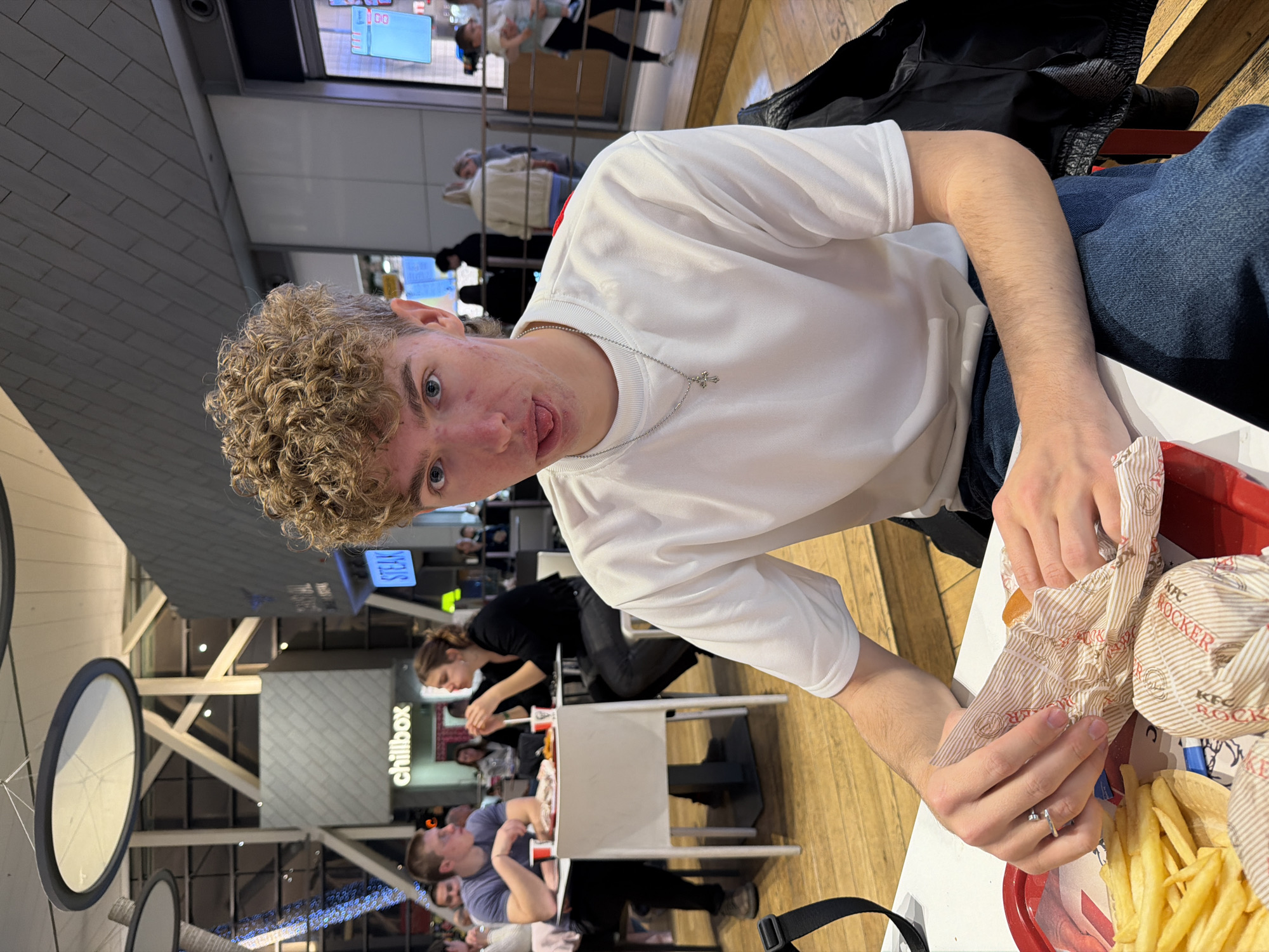 An indoor photograph captures a young man eating at what appears to be a food court or casual restaurant in Sofia, Bulgaria. The image is taken from a slightly tilted, eye-level perspective, focusing on the main subject.
The central figure is a young male with curly, light brown hair and blue eyes, looking directly at the camera with a playful expression, his tongue slightly out. He wears a plain white long-sleeved t-shirt and blue jeans, accessorized with a silver chain featuring a small cross pendant. A silver ring is visible on his left ring finger. He is in the process of unwrapping a food item, likely a burger, held in both hands. A red plastic tray in front of him contains a generous portion of French fries and another wrapped item, which prominently displays "KFC ROCKER" text on its wrapper.
In the background, several other patrons are seated at tables. A woman with dark hair is visible behind the main subject, also eating. The setting features a light-colored table, a dark jacket or bag resting beside the subject, and a wooden or wood-patterned floor. The ceiling reveals modern architectural elements including exposed beams and large circular light fixtures. In the distance, signs for "chillbox" and "STEAK" are visible, indicating other food establishments within the venue. The overall atmosphere suggests a casual dining experience in a busy public space. Artificial lighting illuminates the scene.