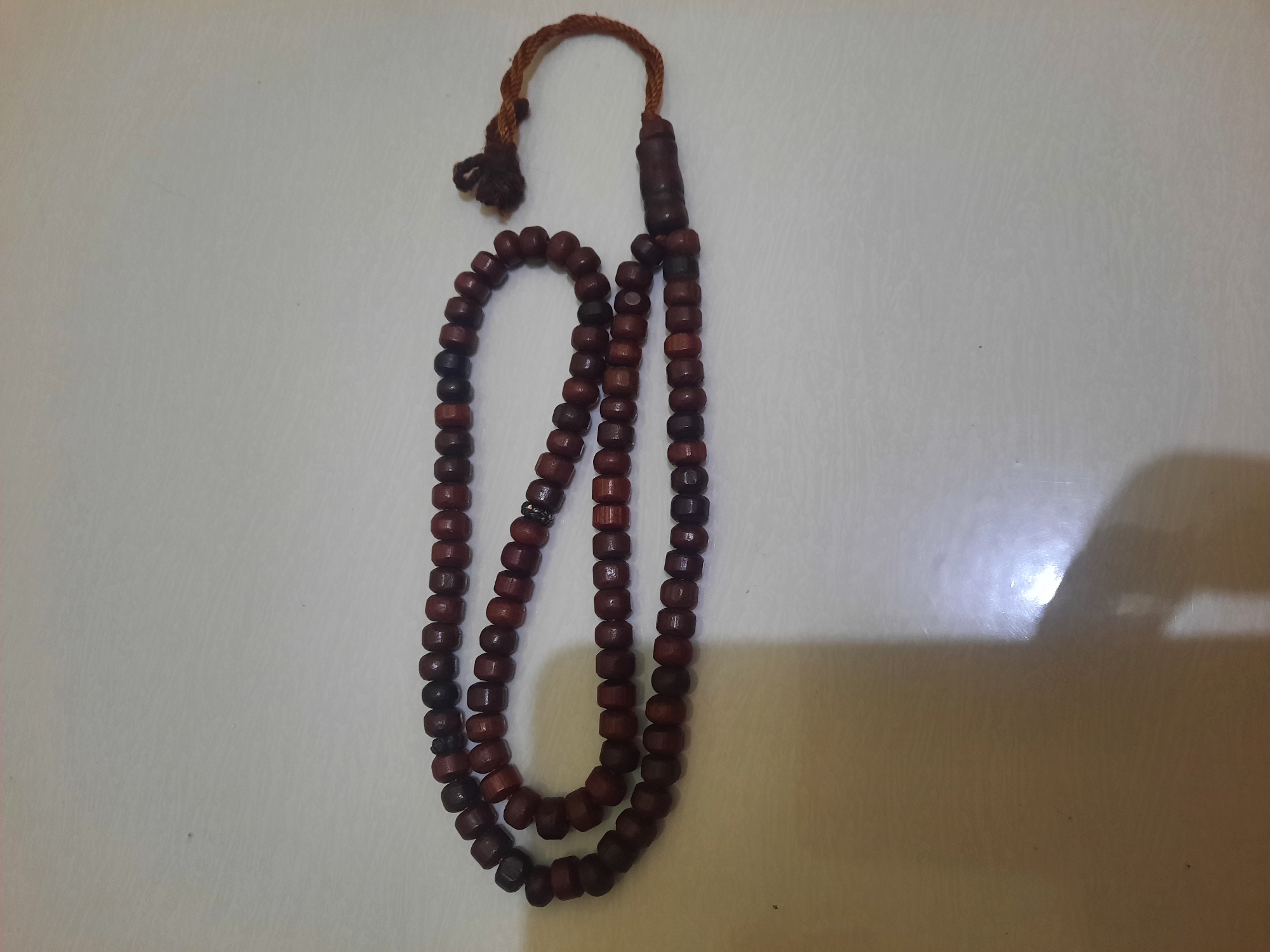 A close-up, overhead shot shows a string of brown wooden prayer beads, also known as tasbih, laid out on a textured, off-white surface. The beads are roughly spherical, with a slight oblong shape and a matte finish. They are strung together with dark thread, and the string forms a loose loop. A small tuft of brown thread and a thicker, carved wooden bead are visible at one end of the string, forming a tassel. The surface appears to be a countertop or table, with subtle variations in texture and tone. Shadows are cast across the surface, with a brighter, almost white area on the right side suggesting a light source. There are no people or animals visible in the image. The image lacks any specific geographic markers that would indicate its location in Lhokseumawe, Indonesia, or any other city.