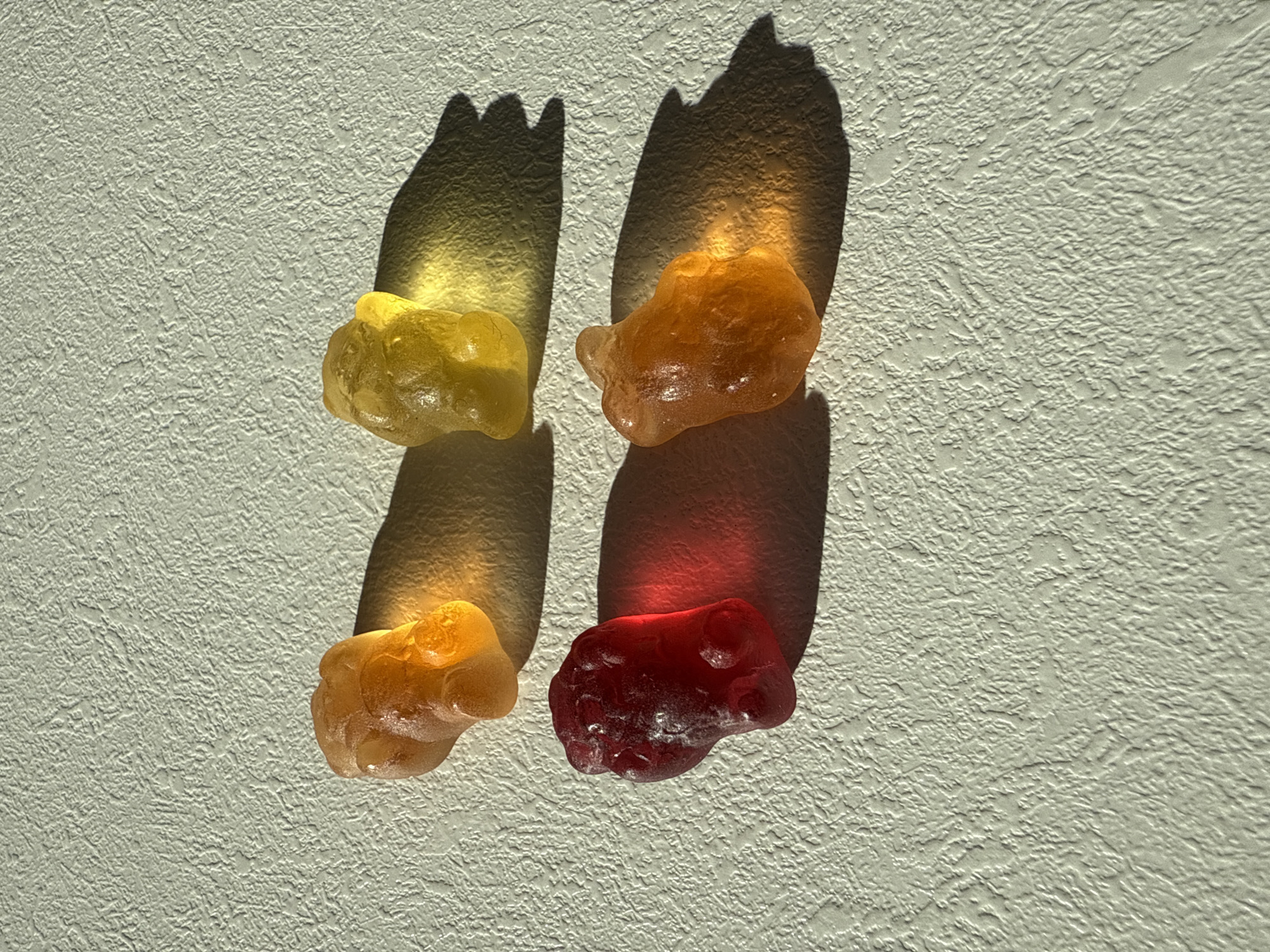 The image shows four gummy bears placed on a textured white surface, likely a wall or a similar flat surface. The gummy bears are of different colors: yellow, orange, orange, and red. Each gummy bear casts a shadow on the surface, which appears distorted due to the irregular shape of the gummy bears and the angle of the light. The lighting suggests a direct light source, possibly sunlight, shining from above. The textured surface adds depth to the image, creating highlights and shadows that accentuate the texture. There is no indication of the location being Votkinsk, Russia, or any other specific location.