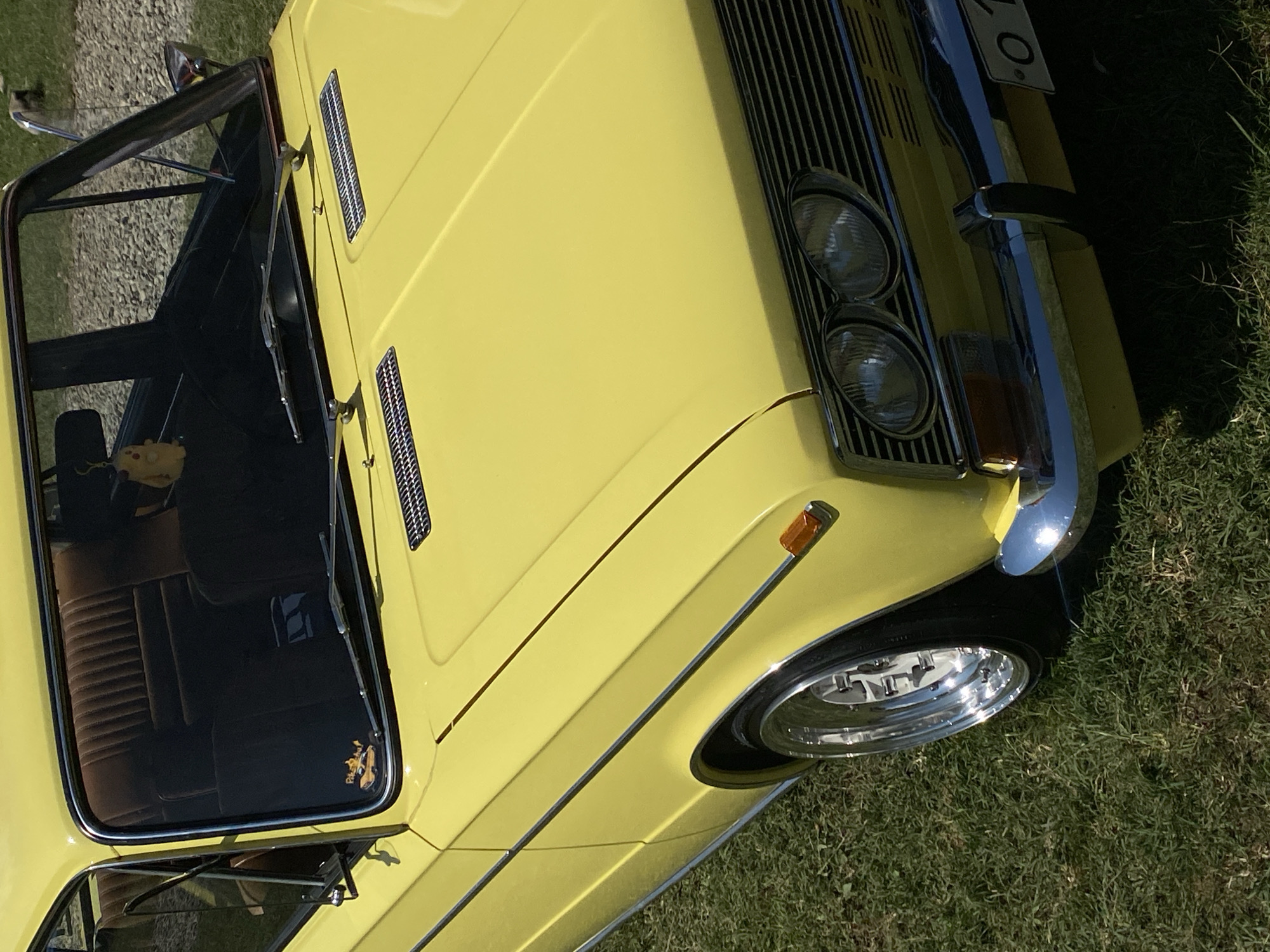 A yellow classic car is parked on a grassy area. The car is a sedan with a chrome bumper and grille. It has two headlights and orange turn signals. The interior of the car is visible through the windshield, showing brown seats. A small yellow Pikachu figure hangs from the rearview mirror, and a sticker of a car with flames is on the lower part of the windshield. The car is parked on a sunny day, with shadows cast on the grass. The location is identified as Bezymyannoe, Russia.