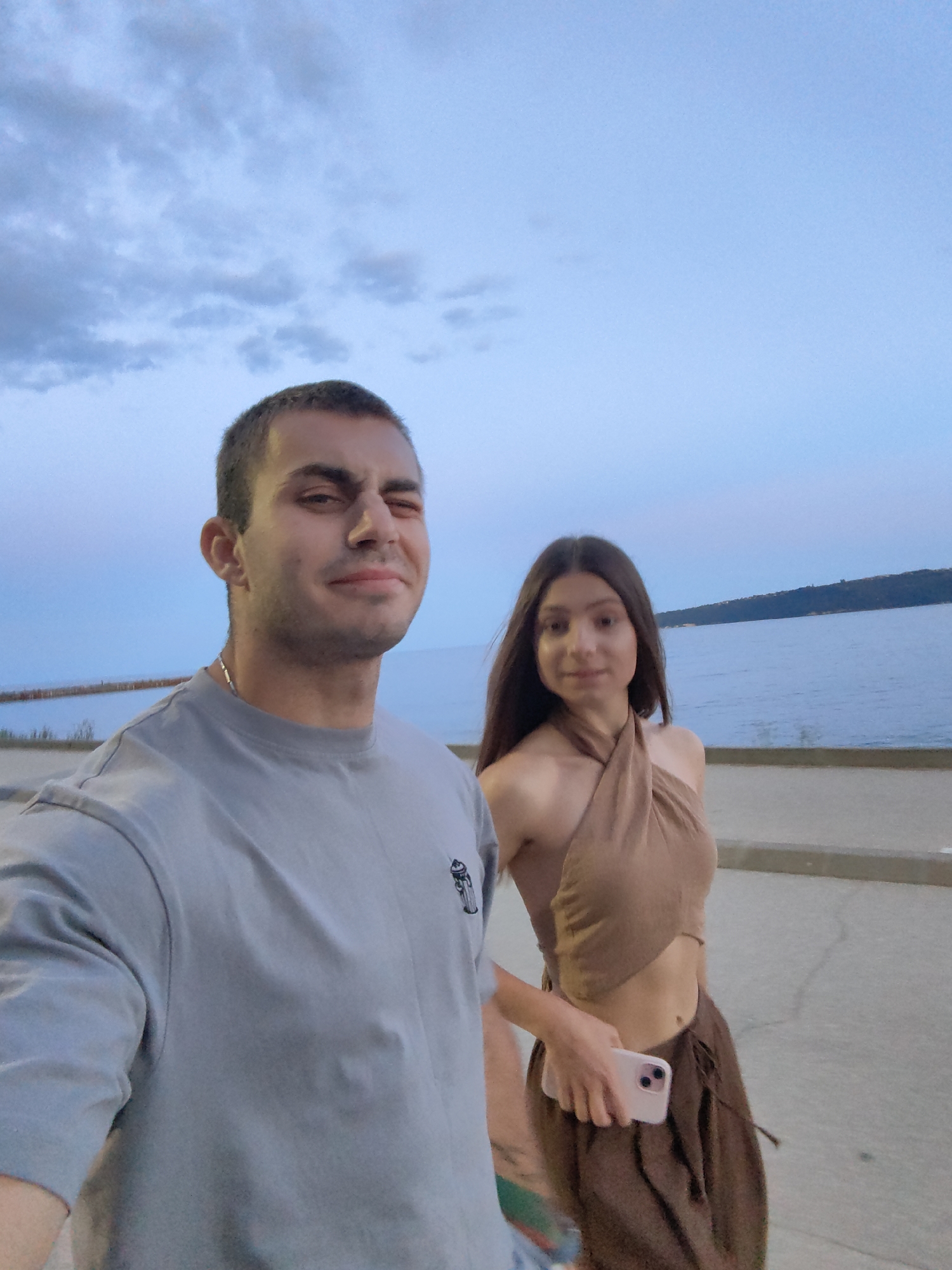 Two individuals, a man and a woman, are walking along a paved promenade by the sea at dusk. The man, in the foreground on the left, is wearing a grey t-shirt and looking towards the camera with a slight squint. The woman, behind him and to the right, is wearing a brown halter top and a brown skirt, holding a pink smartphone. The sea is visible to their right, with a faint coastline and hills in the background. The sky is a dusky blue with scattered clouds. This appears to be the Sealine of Varna, Bulgaria.