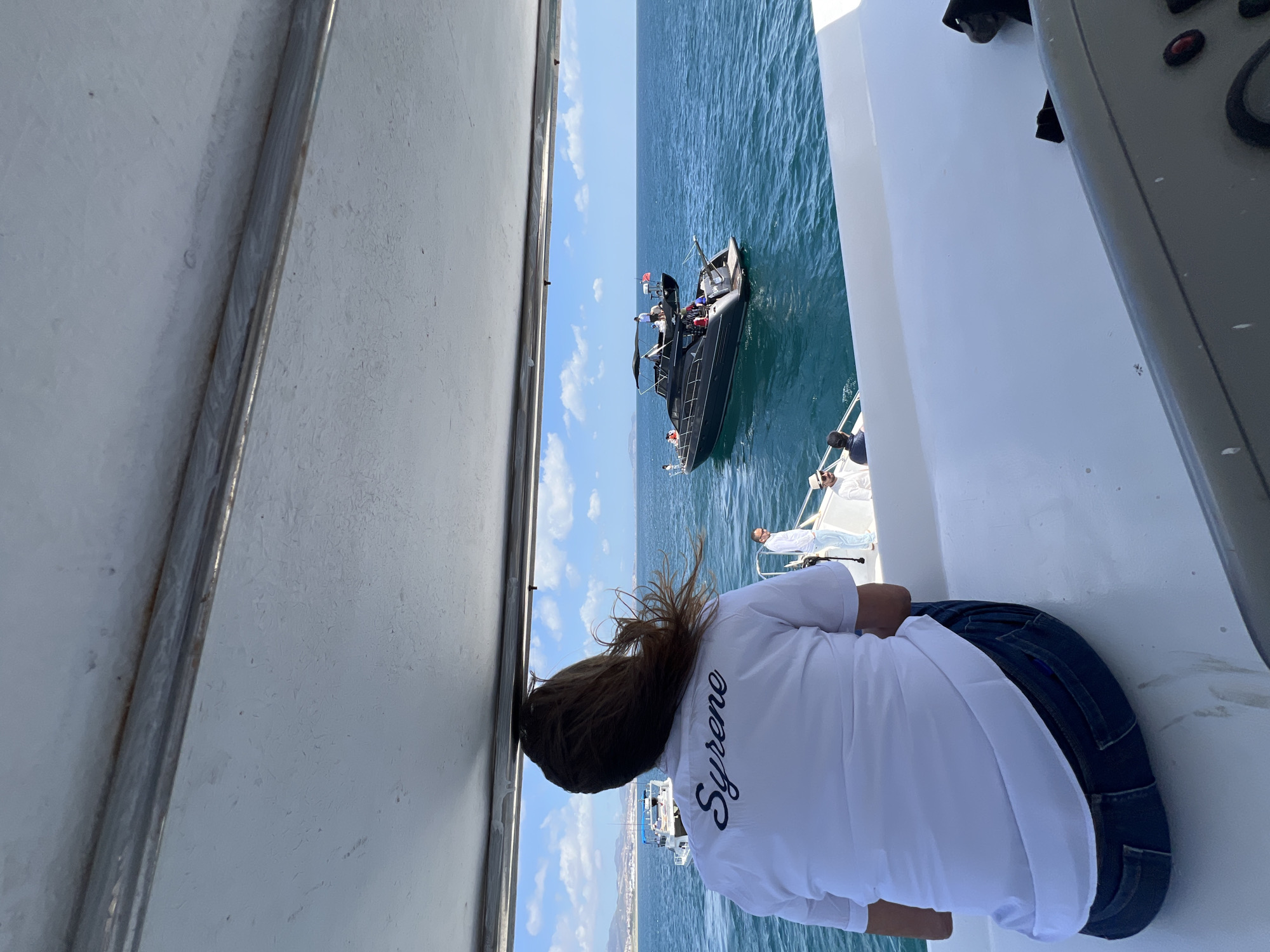 A woman wearing a white shirt with "Syrene" embroidered on the back and blue jeans is seen from behind, looking towards a black boat on the water. The perspective suggests she is on a larger boat or yacht, as indicated by the white structures on either side of the frame. The location is M'Diq, Morocco, as evidenced by the city in the background. A few people are visible on the boat. The sky is blue with scattered clouds.