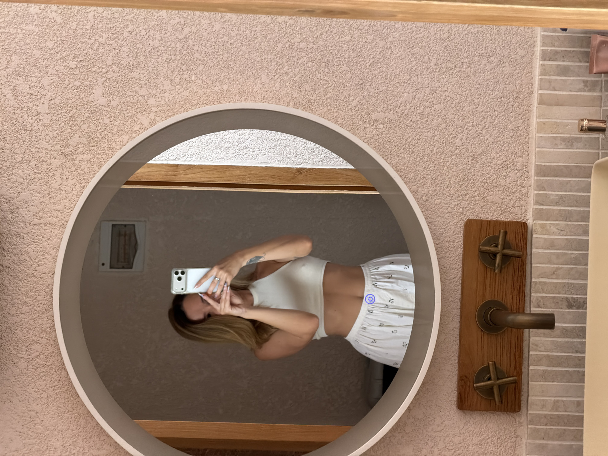 The image is a mirror selfie of a person with blonde hair, wearing a white top and patterned white pajama pants. They are holding a smartphone to take the picture. The mirror is round and framed in light grey, positioned on a textured beige wall. To the right of the mirror is a wooden fixture with a bronze faucet and knobs, set against a backdrop of horizontally-laid stone-like tiles. The reflection within the mirror shows a glimpse of another wall behind the person, with a framed square object visible. The location, Mengwi, Indonesia is not discernible from the visual data in the image.